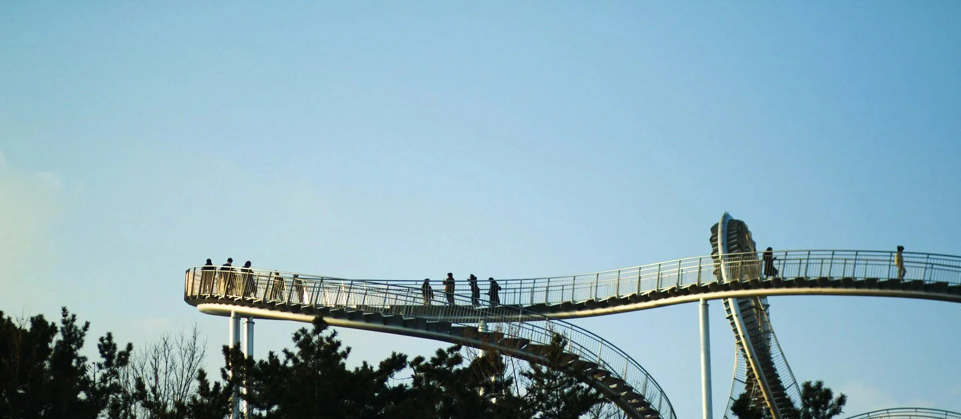 A Ferris wheel at a carnival, seen from below, with people walking or standing on a walkway at the top. The sky is light blue, and there are trees in the foreground.