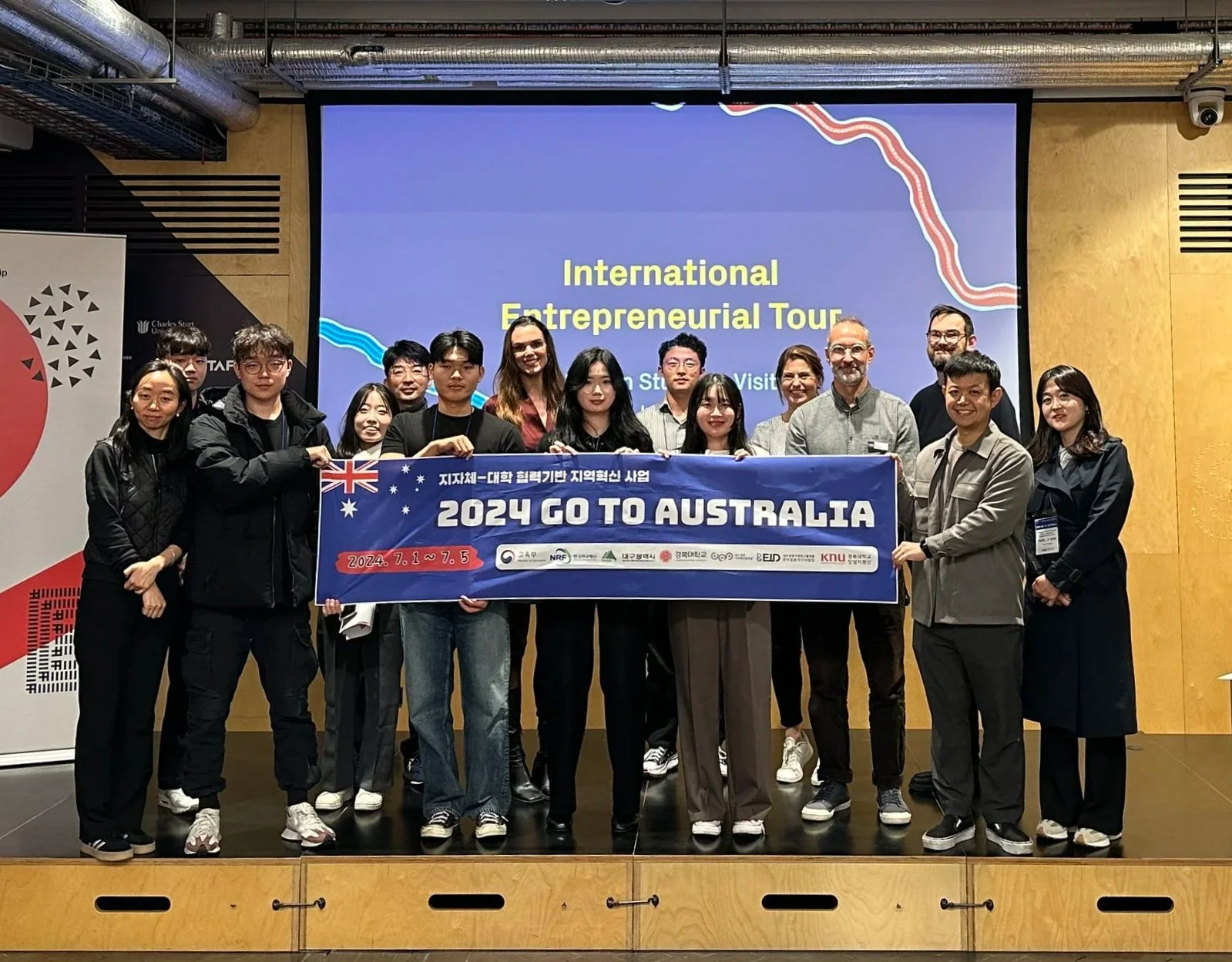 Group of young people and adults posing with a banner announcing a trip to Australia in 2024, in a conference room.