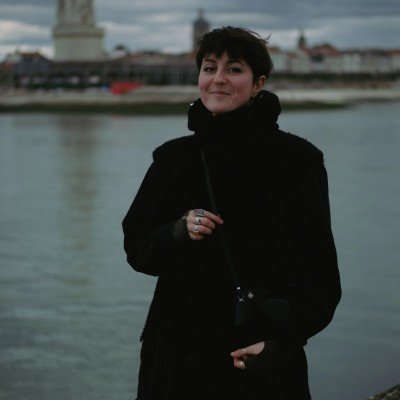 A woman with short hair, wearing a black coat, standing near a river or a port, with a building and statues in the background.