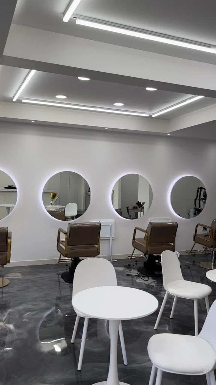 A modern hair salon with brown makeup chairs and round mirrors with LED lighting in a white and gray interior.