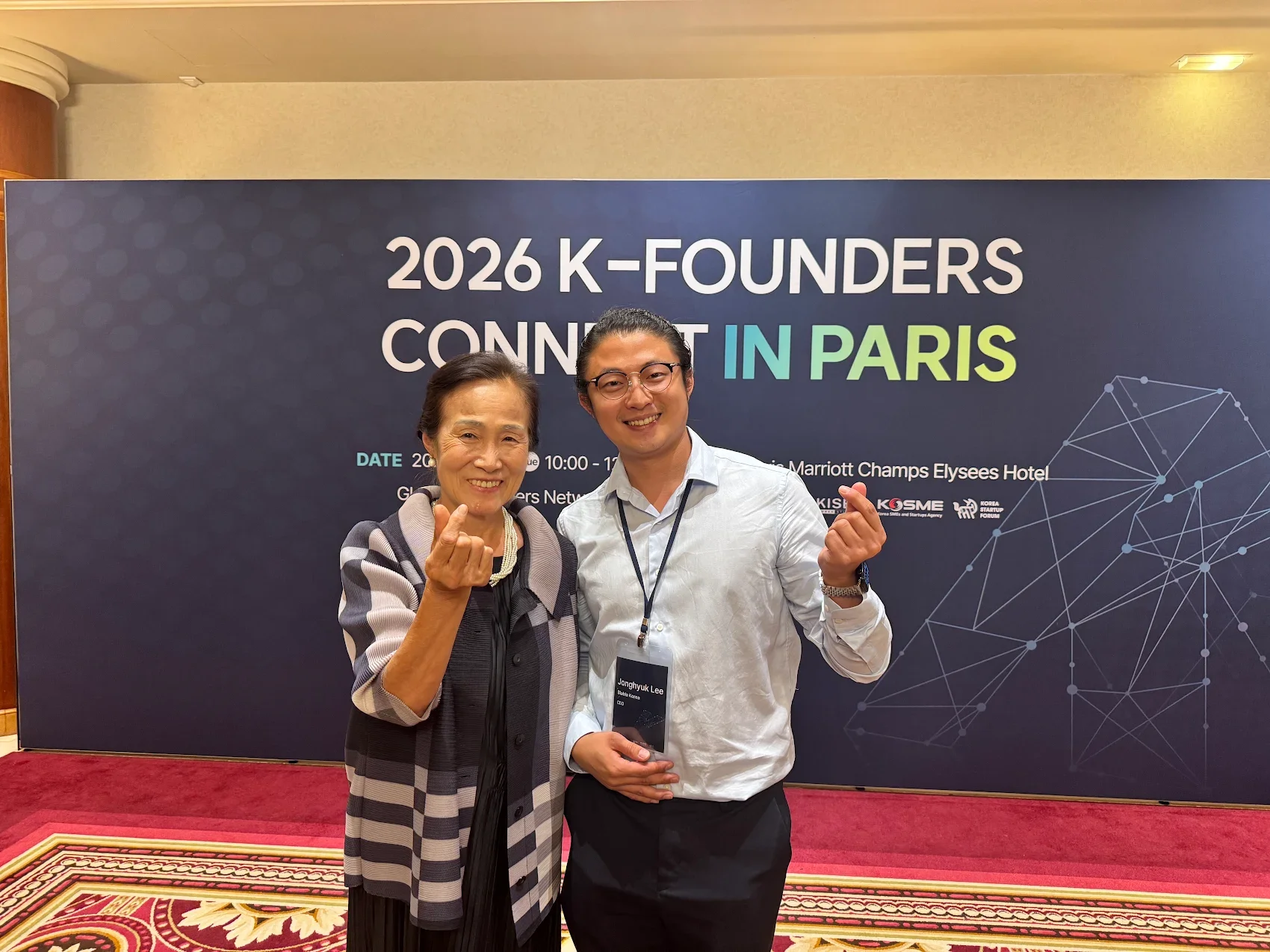 Jonghyuk et la présidente de Kimchi Château posent pour une photo lors d'une conférence à Paris. Elles font un geste de cœur avec leur main, et la femme porte un manteau à carreaux. Le jeune homme porte une chemise blanche et un badge autour du cou.