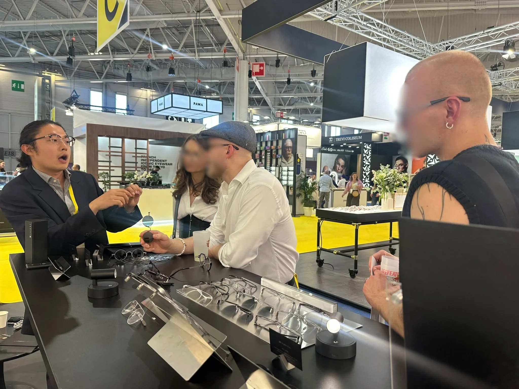 Groupe de personnes discutant autour d'un stand de vente de lunettes dans un centre d'exposition ou un salon. Plusieurs lunettes sont exposées sur le comptoir et deux hommes, dont l'un porte des lunettes, sont engagés dans une conversation.