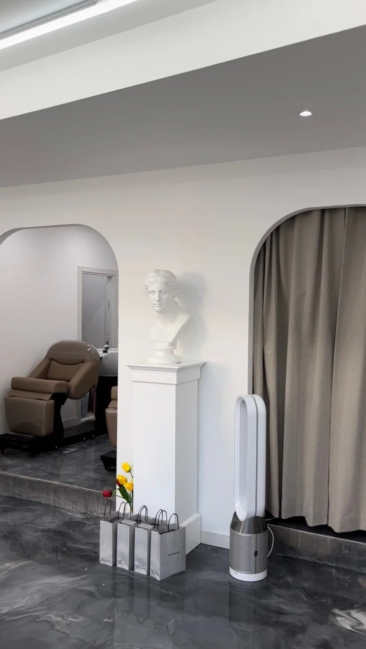 A modern interior featuring a bust sculpture next to a brown leather armchair, a plant with yellow flowers, white shopping bags, an air purifier, and beige curtains in front of an archway.