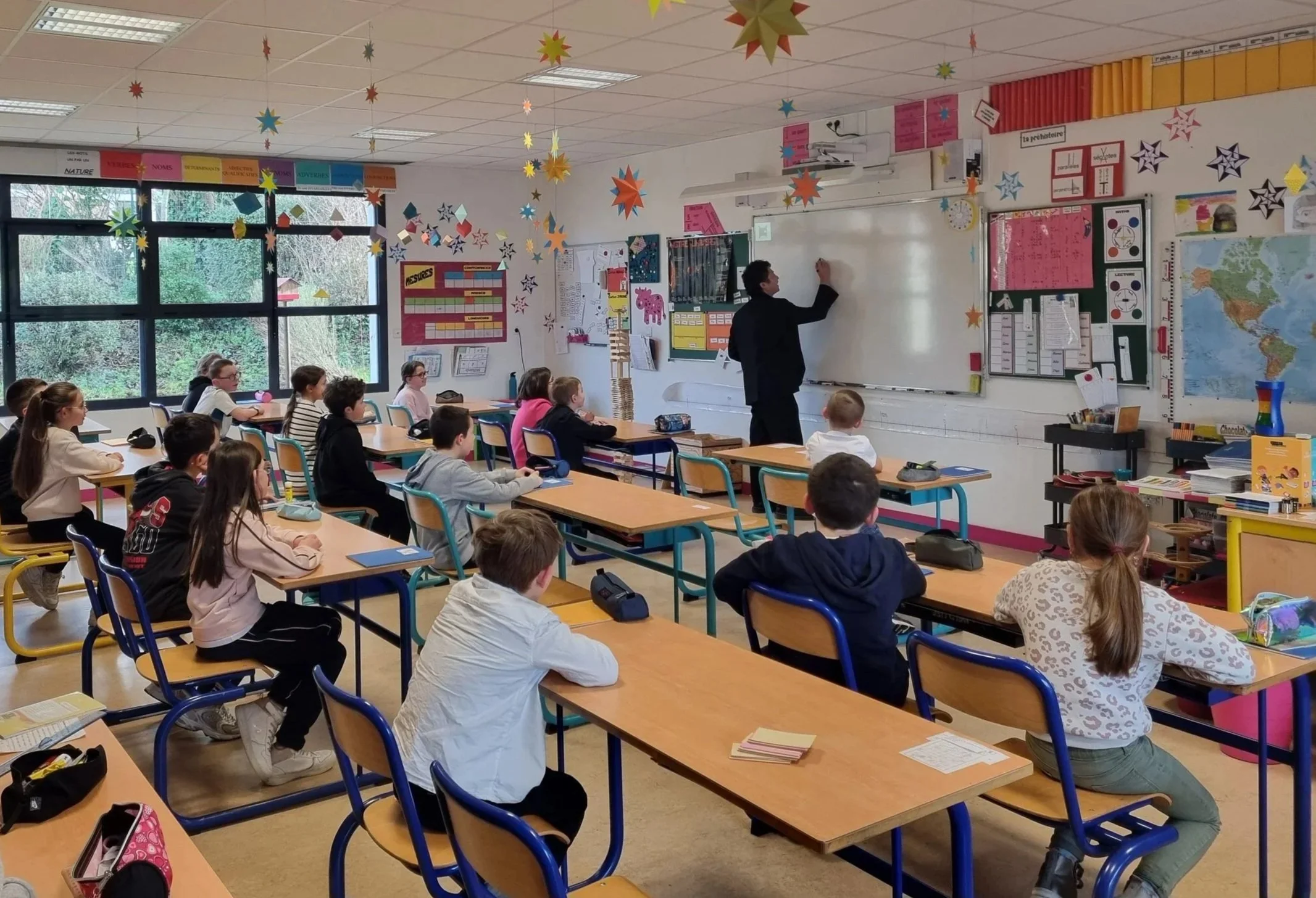 A classroom with students sitting at their desks, a teacher at the front giving explanations. The classroom is decorated with stars hanging from the ceiling, colorful posters, a map, and windows letting in natural light.