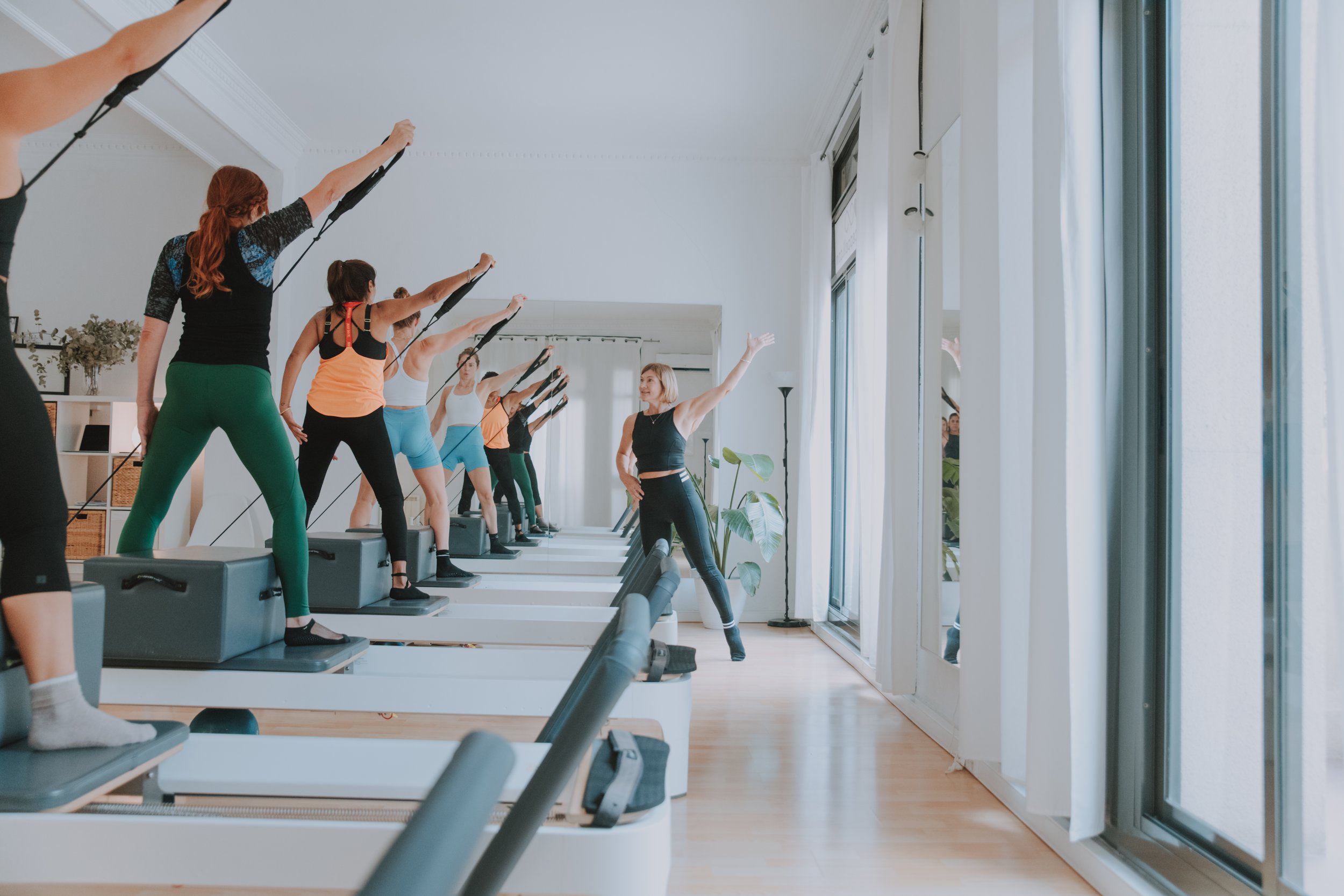 People participating in a fitness class on step platforms in a bright room with large windows, led by an instructor.