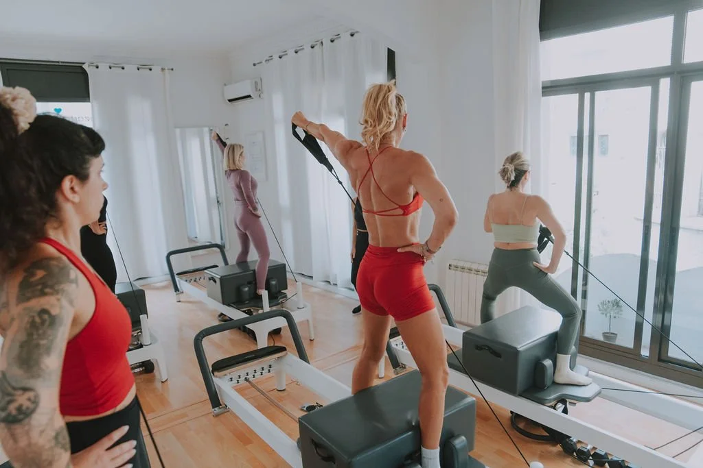 Women participating in a Pilates class using reformer machines in a bright studio with large windows and white curtains.