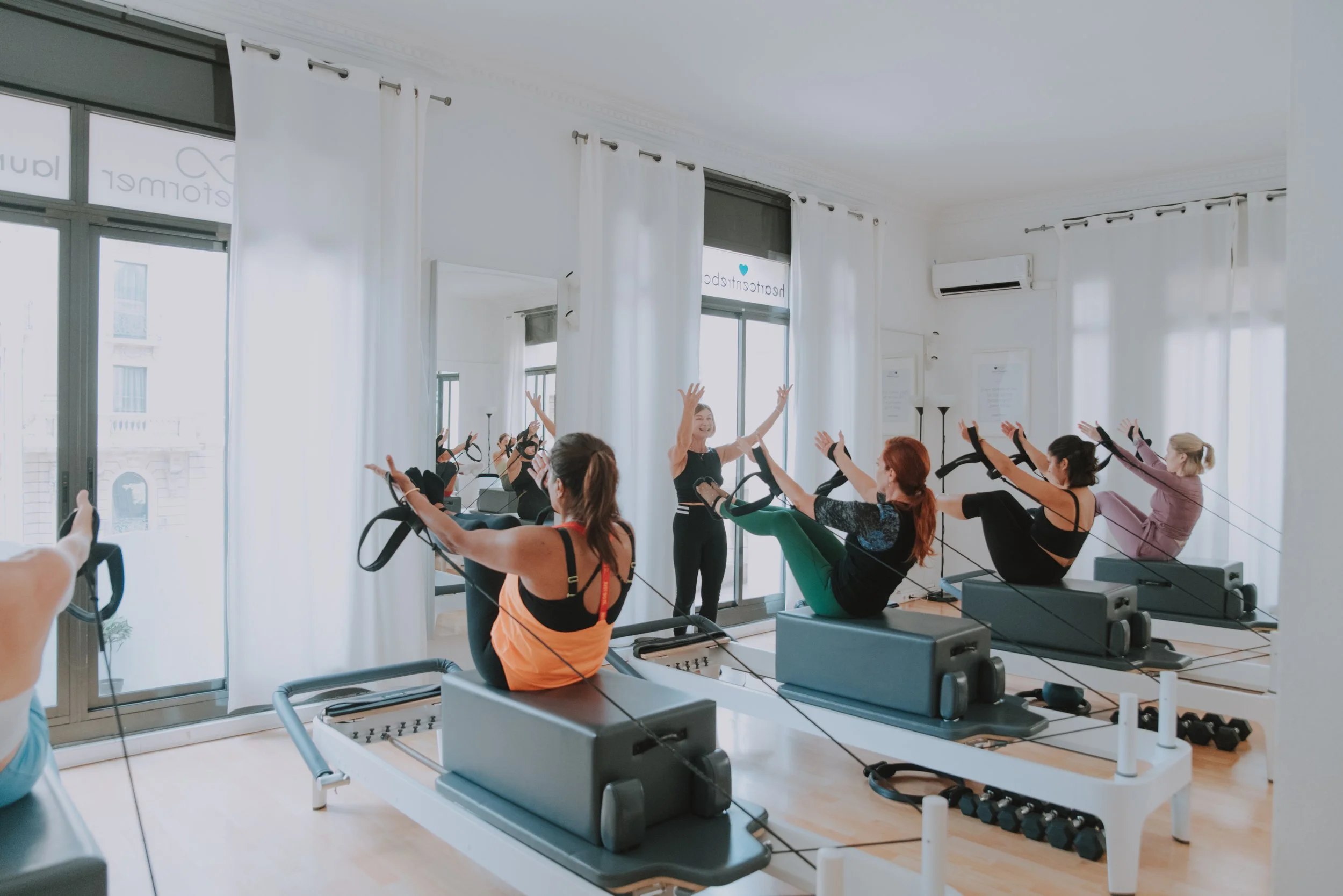 Group of women participating in a Pilates class using reformer machines in a bright studio with large windows and white curtains.