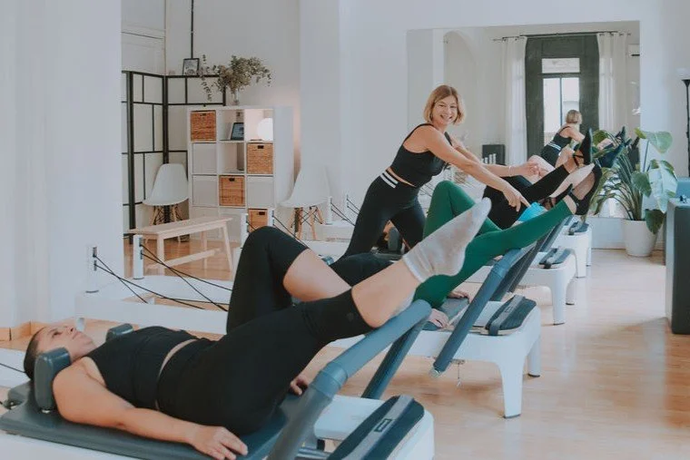 Women exercising on Pilates reformers in a bright, modern room with plants and shelves.