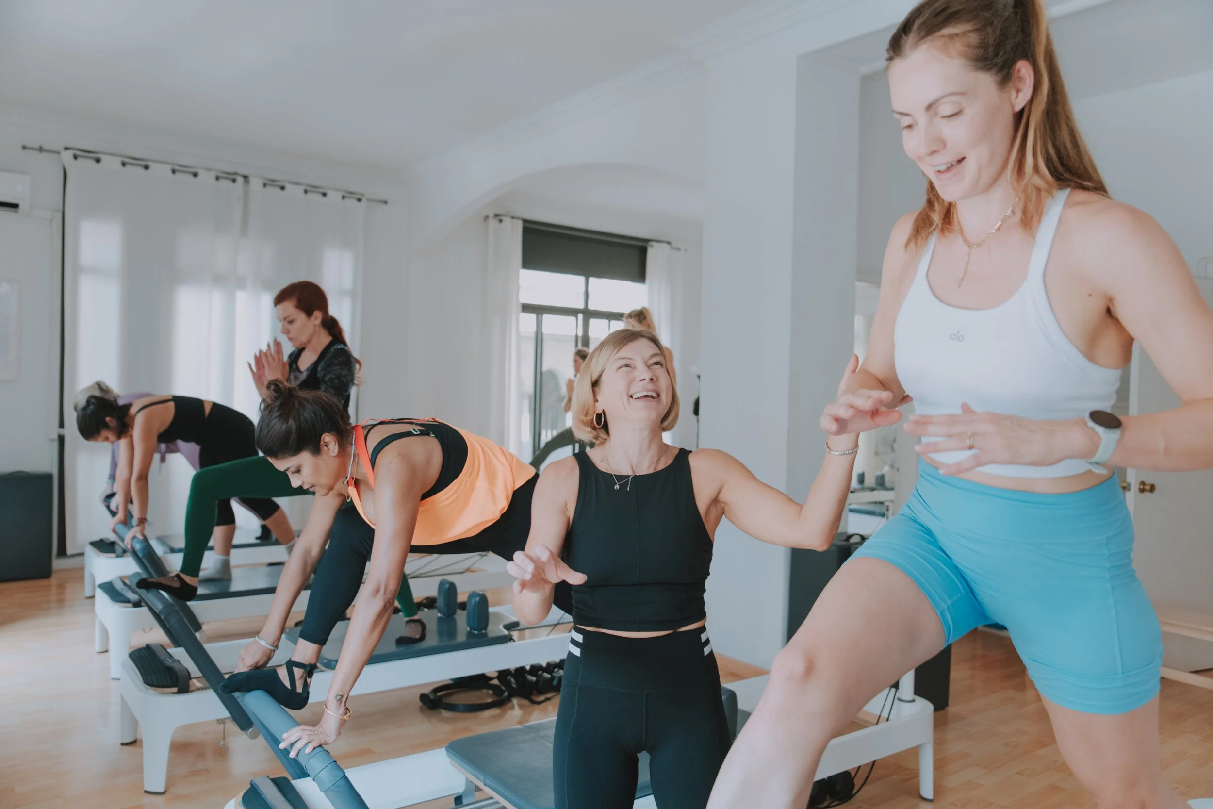 A group fitness class in a bright room with large windows, where women are using steps for an aerobics workout. One woman is guiding, smiling, and talking to another woman who is smiling back.
