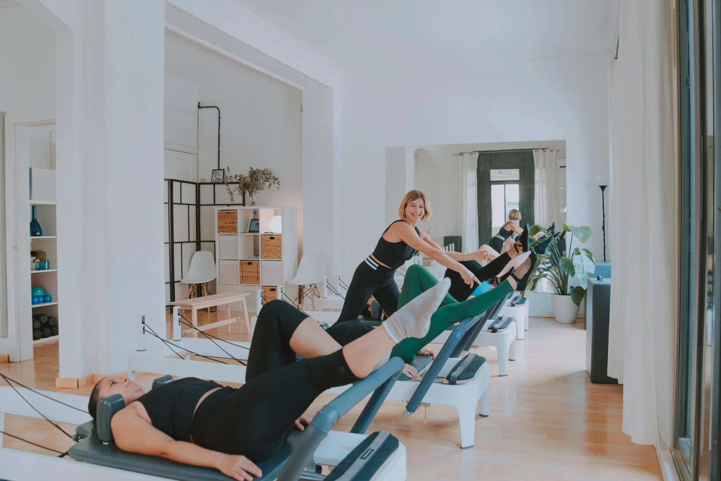 Women exercise on reformer Pilates machines in bright room with large windows, plants, and modern decor.