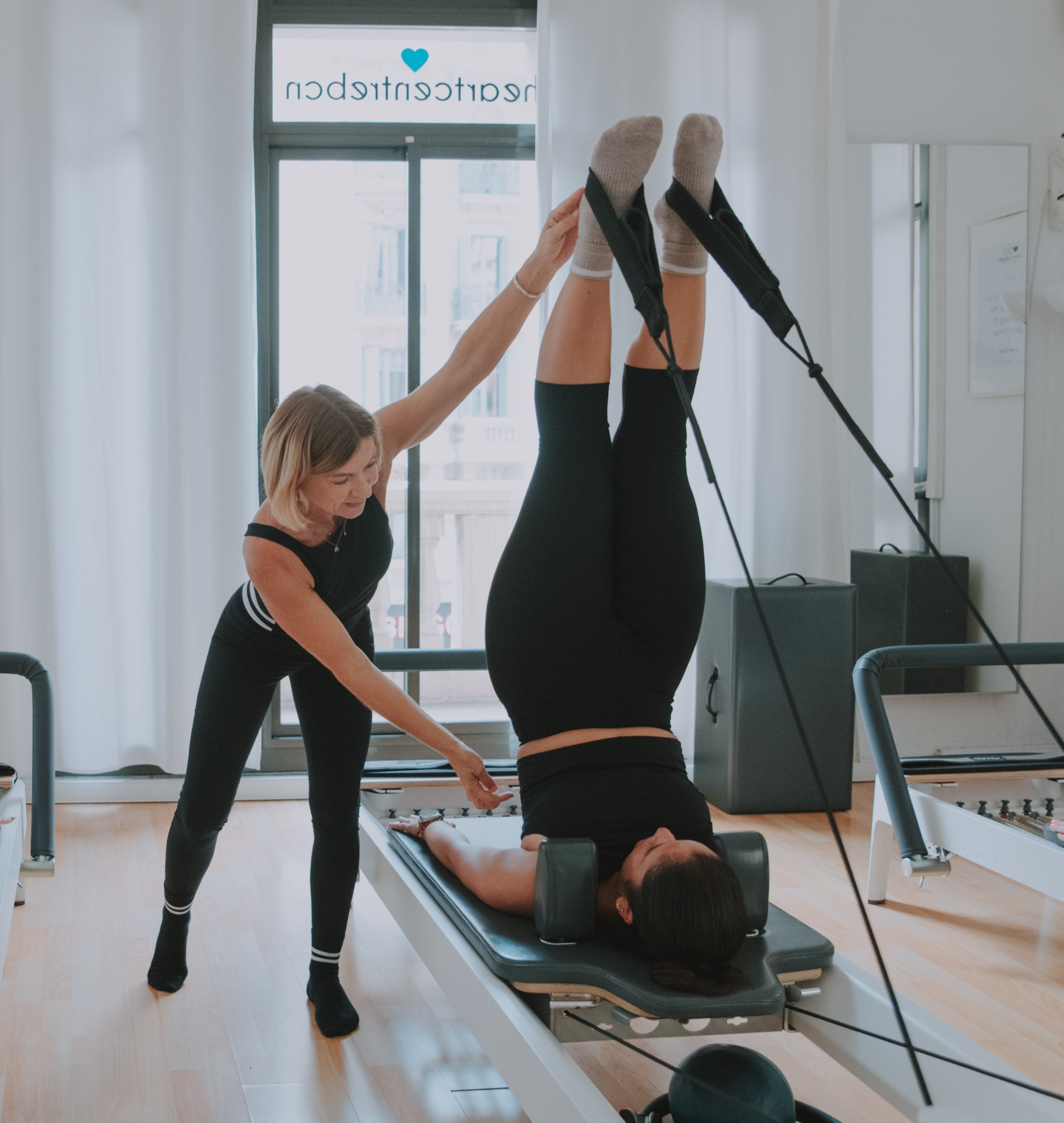 A physical therapist assisting a woman with an exercise on a therapy table, with a large window and a logo with a blue heart in the background.