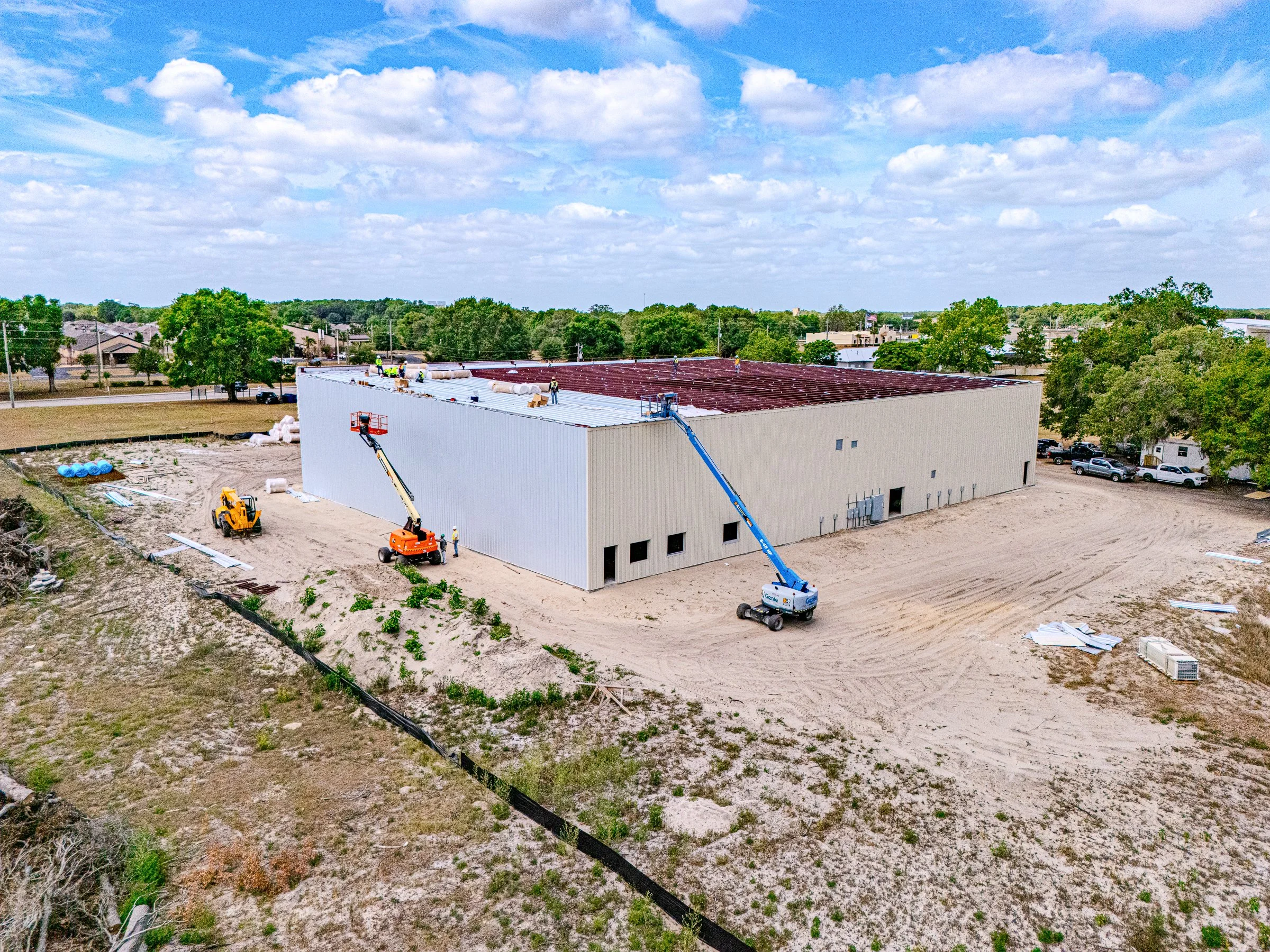 Construction site with workers installing a roof on a large building. The building has white walls, and the roof is covered with red panels. Construction equipment, including a cherry picker and a small excavator, are visible. There are trees and a residential area in the background, under a partly cloudy blue sky.