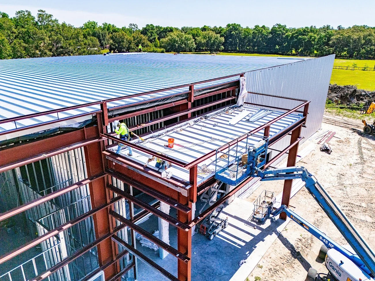 Construction workers installing a metal roof on a building with a steel framework, using a lift and scaffolding in a rural area.