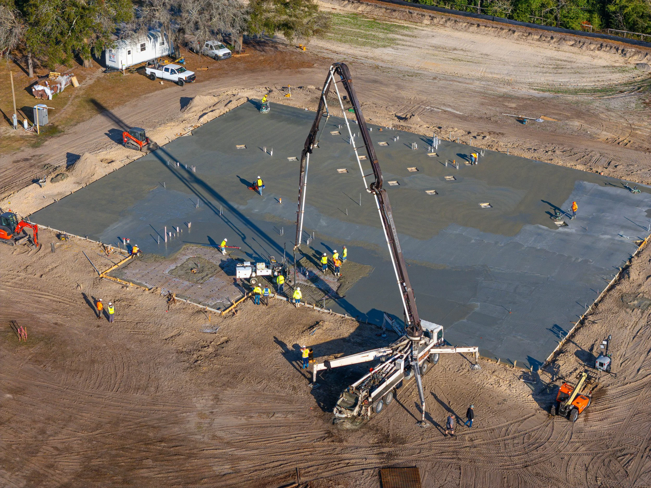 An aerial view of a construction site with workers pouring concrete for a building foundation, surrounded by dirt, construction vehicles, and equipment.