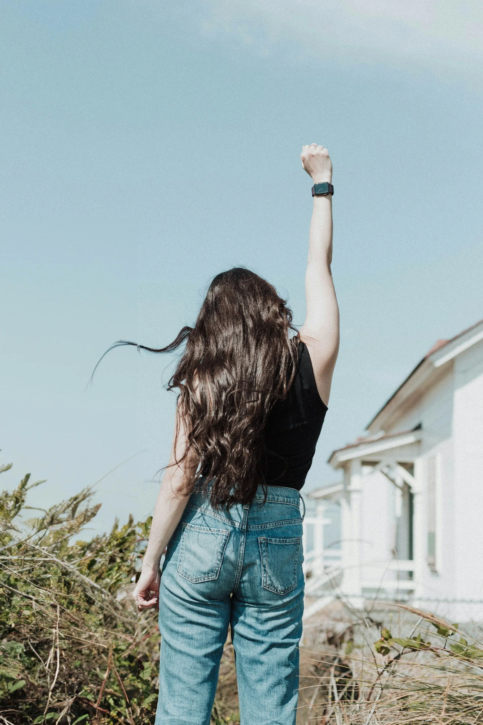 A woman with long wavy hair wearing a black top and blue jeans, raising her right arm with a smartwatch in front of a background of green shrubs and a row of white houses with porches under a clear blue sky.