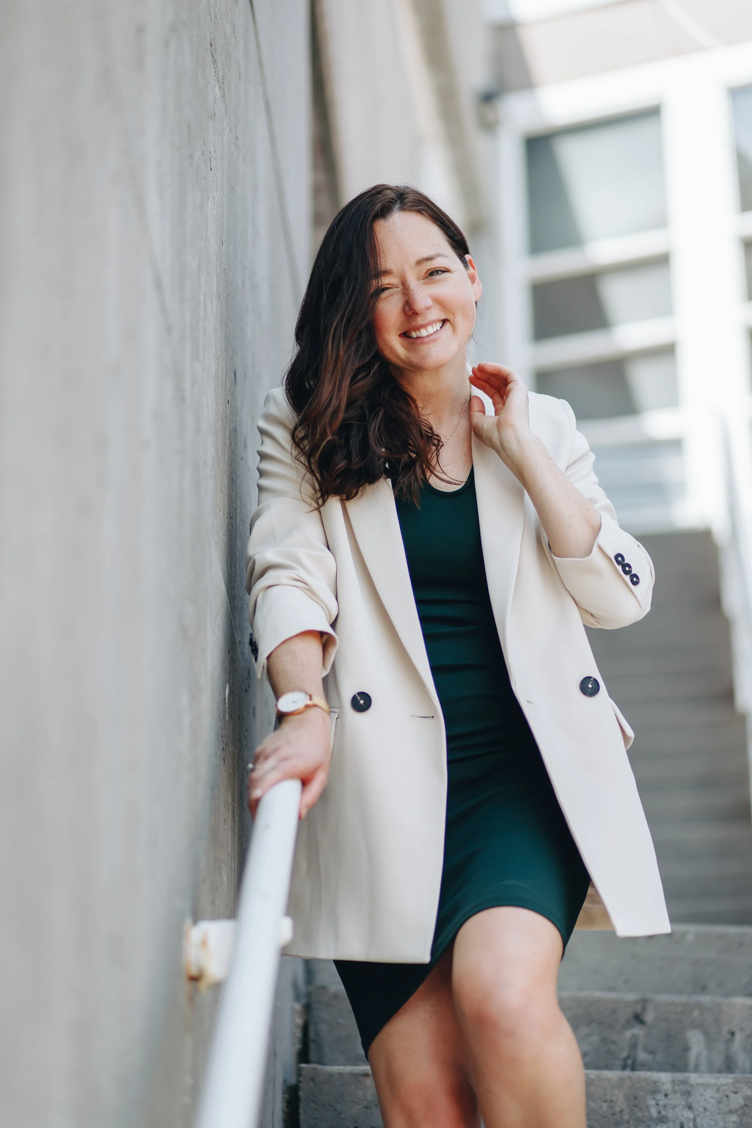 A woman with shoulder-length dark hair, smiling and wearing a white blazer over a dark dress, standing on outdoor stairs next to a concrete wall.