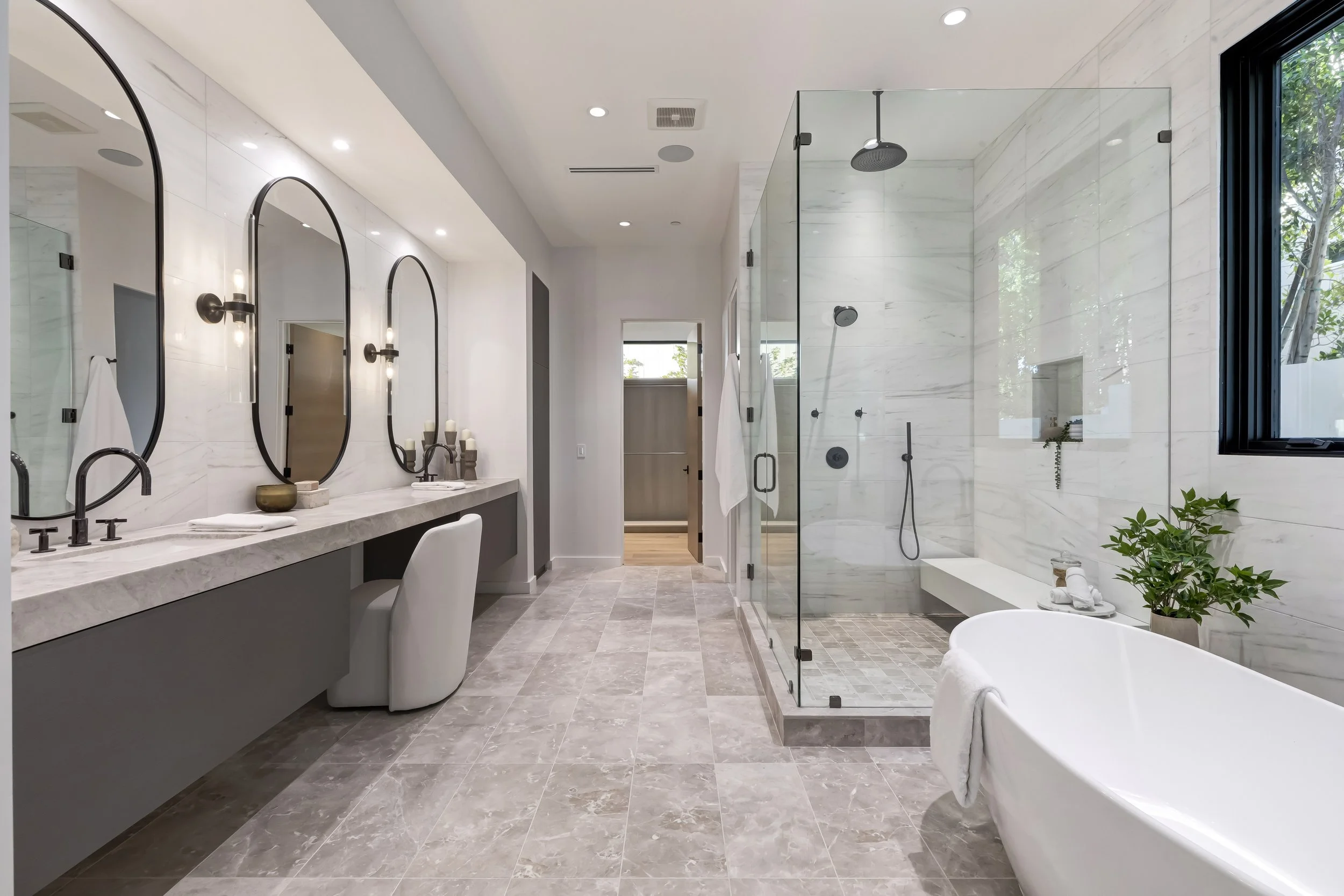 Modern bathroom with a large walk-in shower with glass enclosure, a freestanding bathtub, a double vanity with oval mirrors, and beige tiled floors and walls, illuminated by recessed ceiling lights and natural light from a window.