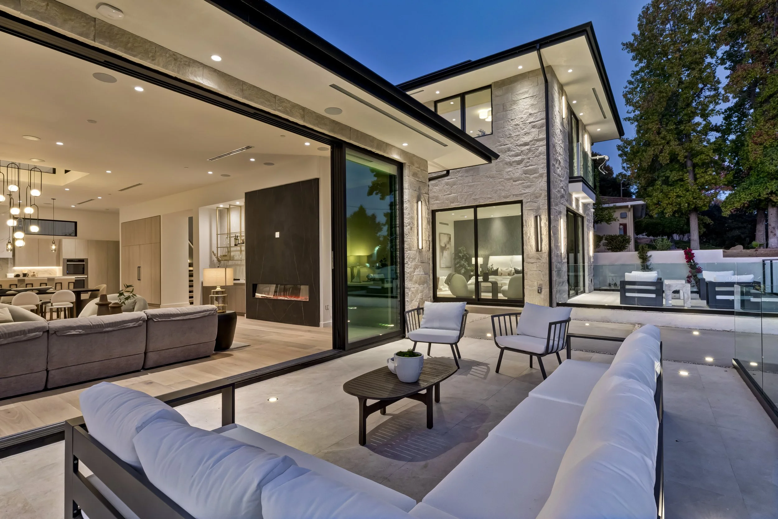 [hardscaping] Open sliding glass walls connecting the great room to a covered patio at twilight, Encino custom build