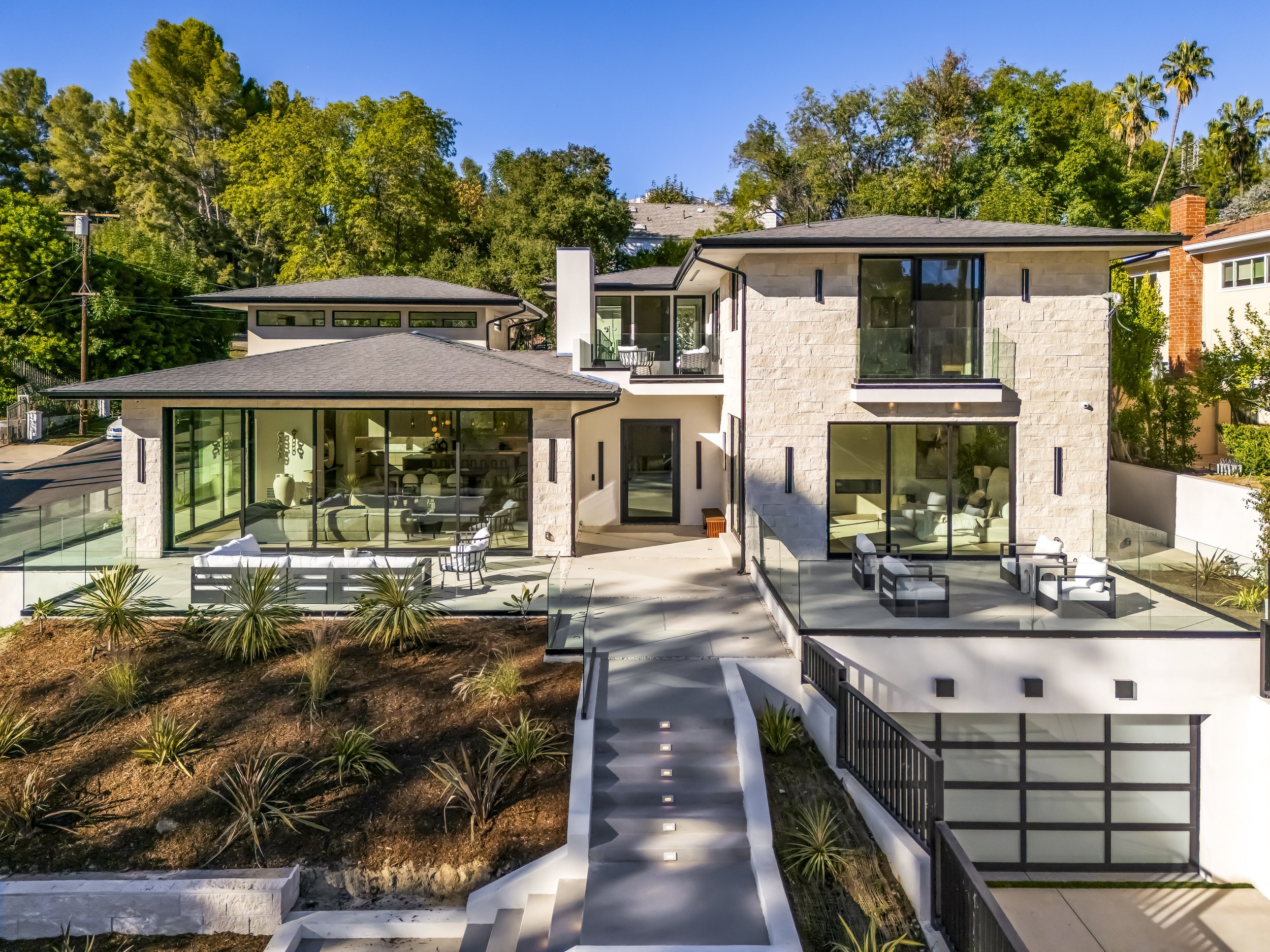 Modern two-story house with large glass windows and balcony, surrounded by trees and landscaped yard, daylight, clear blue sky.