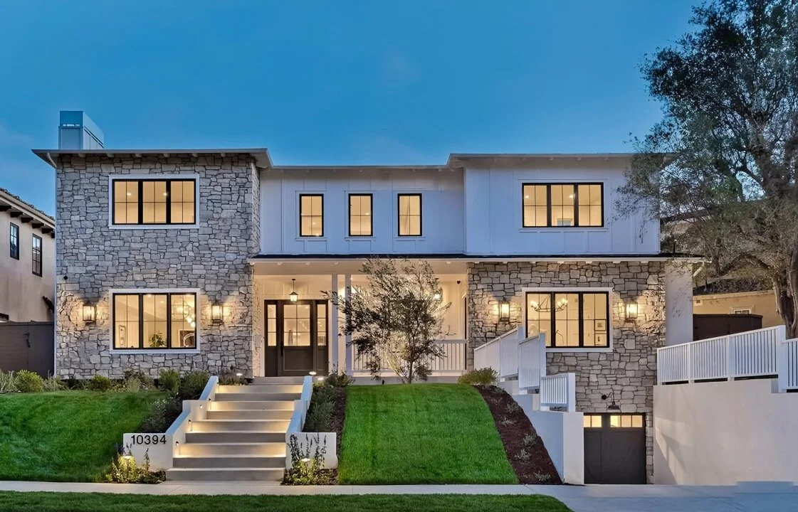 Modern two-story house with stone and white siding exterior, illuminated windows, well-lit front steps, and a landscaped yard at dusk.