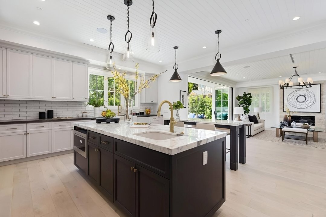 Modern kitchen with white cabinets, black island with marble countertop, pendant lighting, and open living area with sofas, large windows, and artwork.