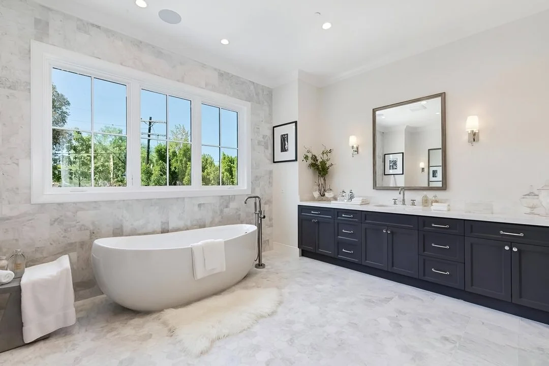 Modern bathroom with white freestanding bathtub, large window showing greenery outside, and a navy blue vanity with a large mirror. White walls and marble flooring.
