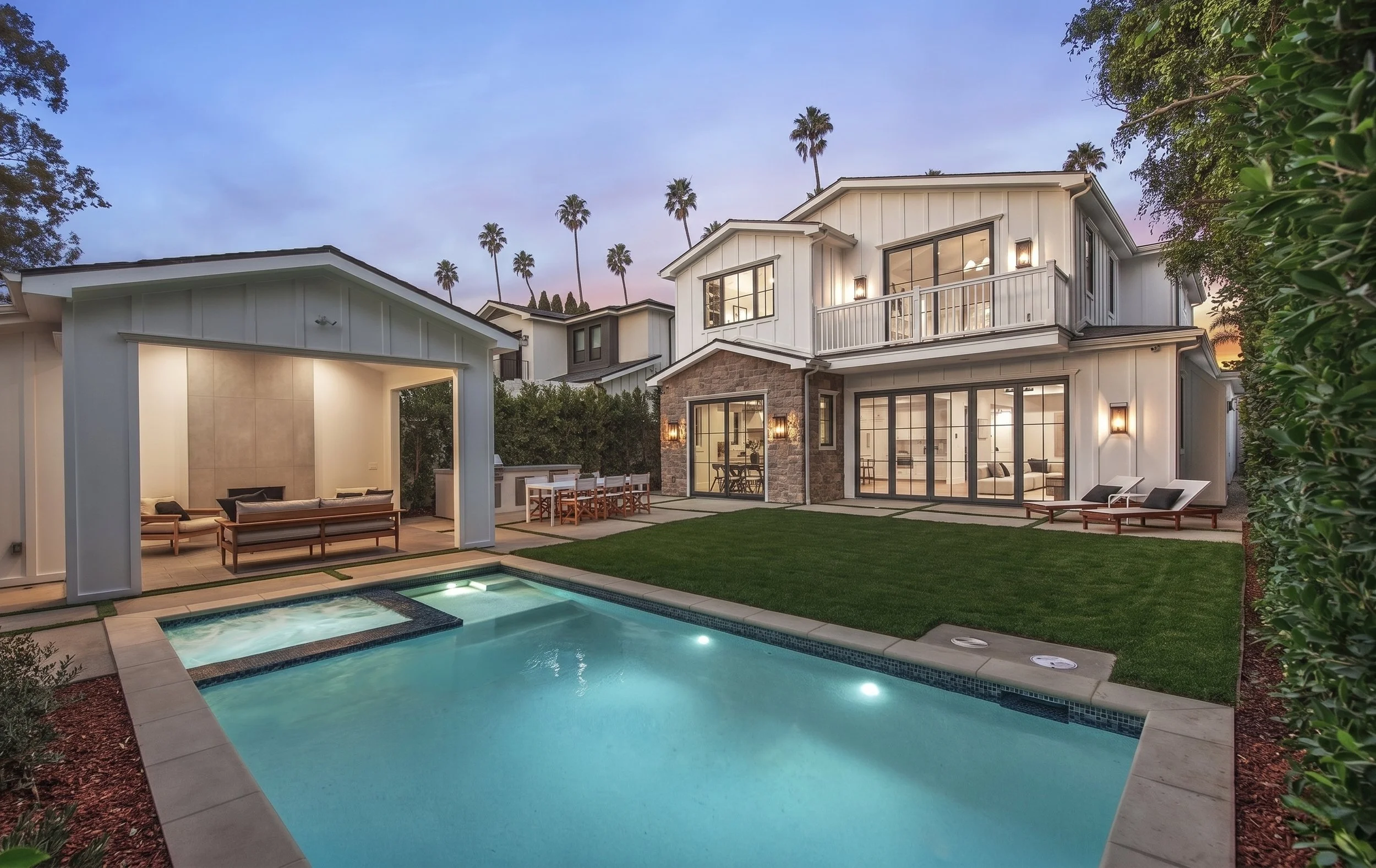 A modern residential house with a driveway, landscaped front yard with drought-tolerant plants, small rocks, and mulch, and tall trees including palm trees, during sunset.