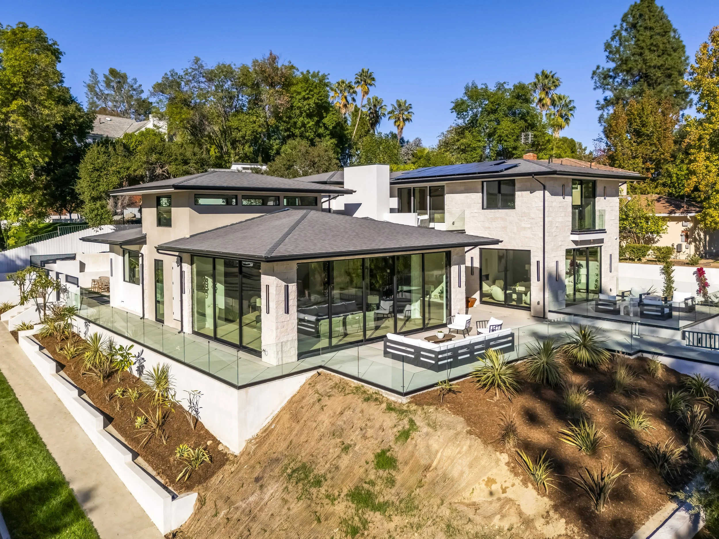 Aerial view of a modern house with a backyard, surrounded by trees and neighboring homes, under a colorful sunset sky with mountains in the background.