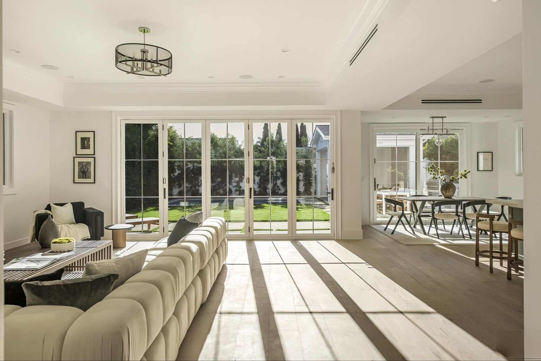 Bright living room with white walls and large glass sliding doors leading to a backyard with green grass and trees. There is a beige tufted sofa with a black armchair next to it, a wooden coffee table with decorative items, and framed wall art. To the right, a dining area has a round table with four chairs and a large decorative vase with greenery. Natural sunlight streams through the windows, casting shadows on the wooden floors.