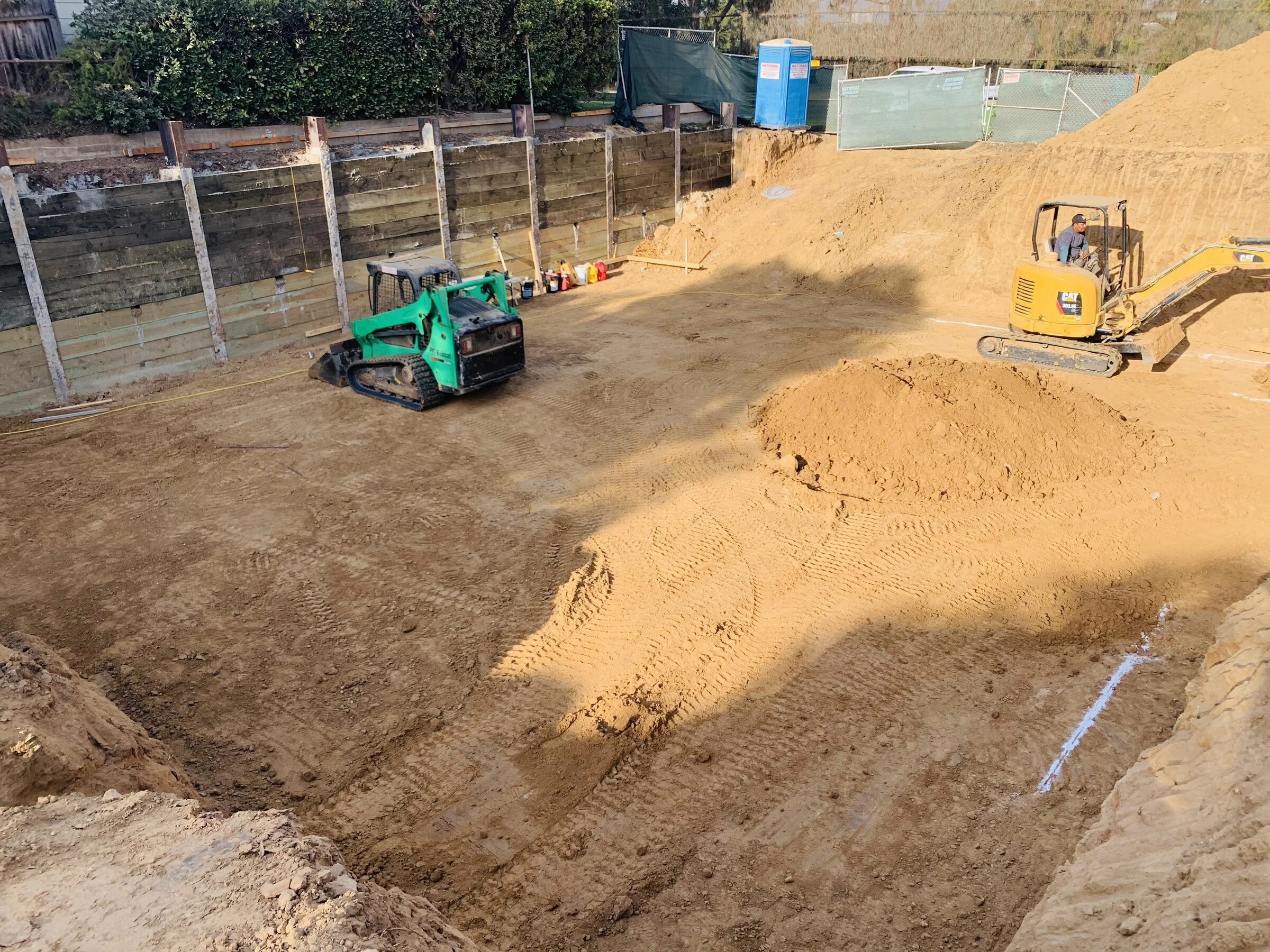 Construction site with excavated dirt and heavy machinery, including a small yellow excavator and a green compact loader, with plywood retaining wall and fencing in the background.