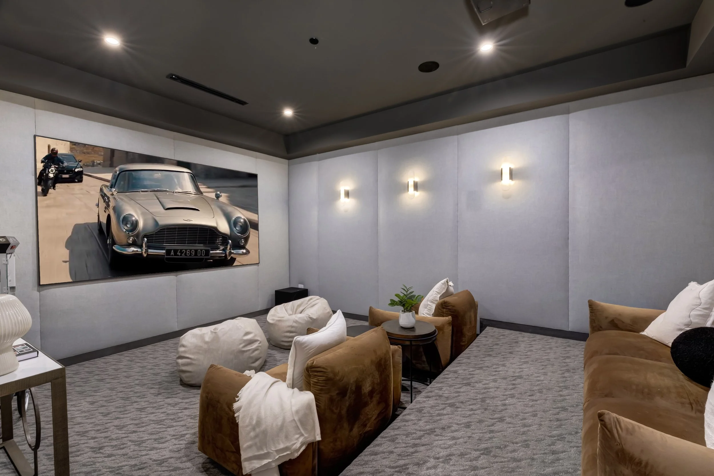 A cozy living room with beige armchairs and pillows, a large wall art of a vintage silver car, and modern wall lights.