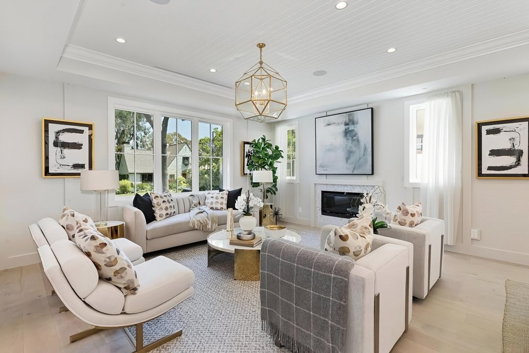 Bright living room with white walls, multiple windows, and contemporary furniture including sofas with patterned and solid pillows, a round coffee table, and abstract artwork on the walls. There's a chandelier hanging from the ceiling and a fireplace beneath a large abstract painting.
