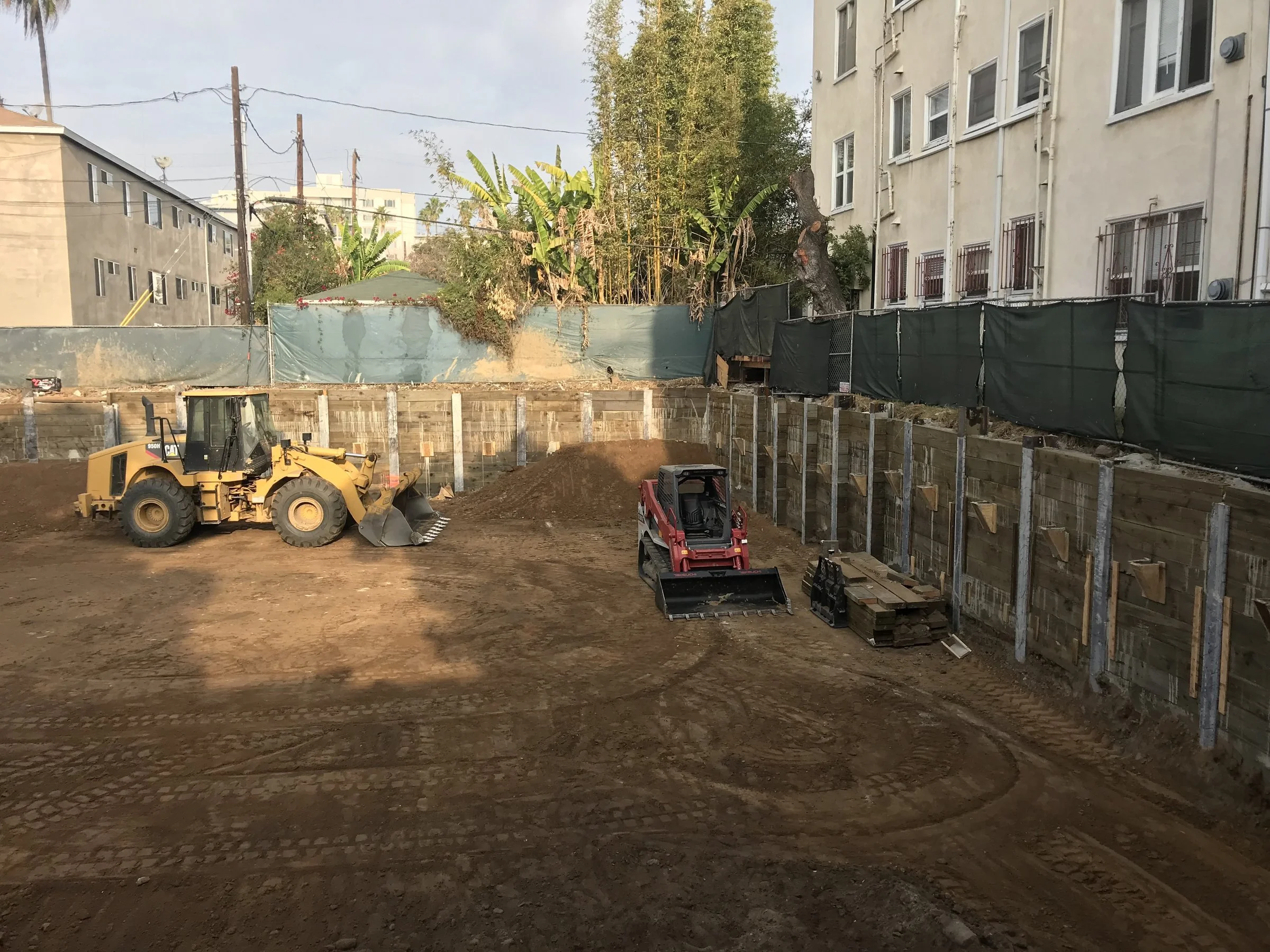 [new-construction] Wide view of a shored basement excavation with timber lagging and earthmoving equipment in Santa Monica