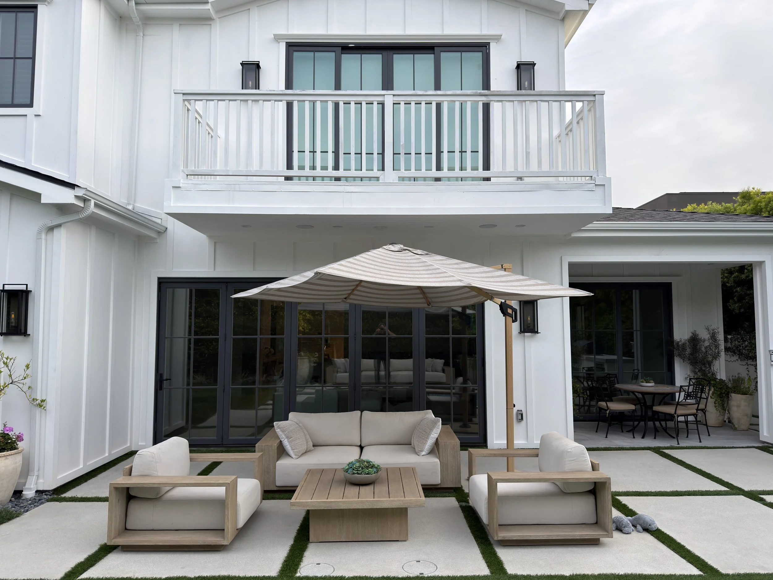 Outdoor patio area with beige cushioned chairs, a sofa, and a wooden coffee table under a large patio umbrella. The area is covered with concrete pavers separated by strips of green grass, and the house has large glass doors and windows with black frames, and a white exterior with a balcony on the upper level.
