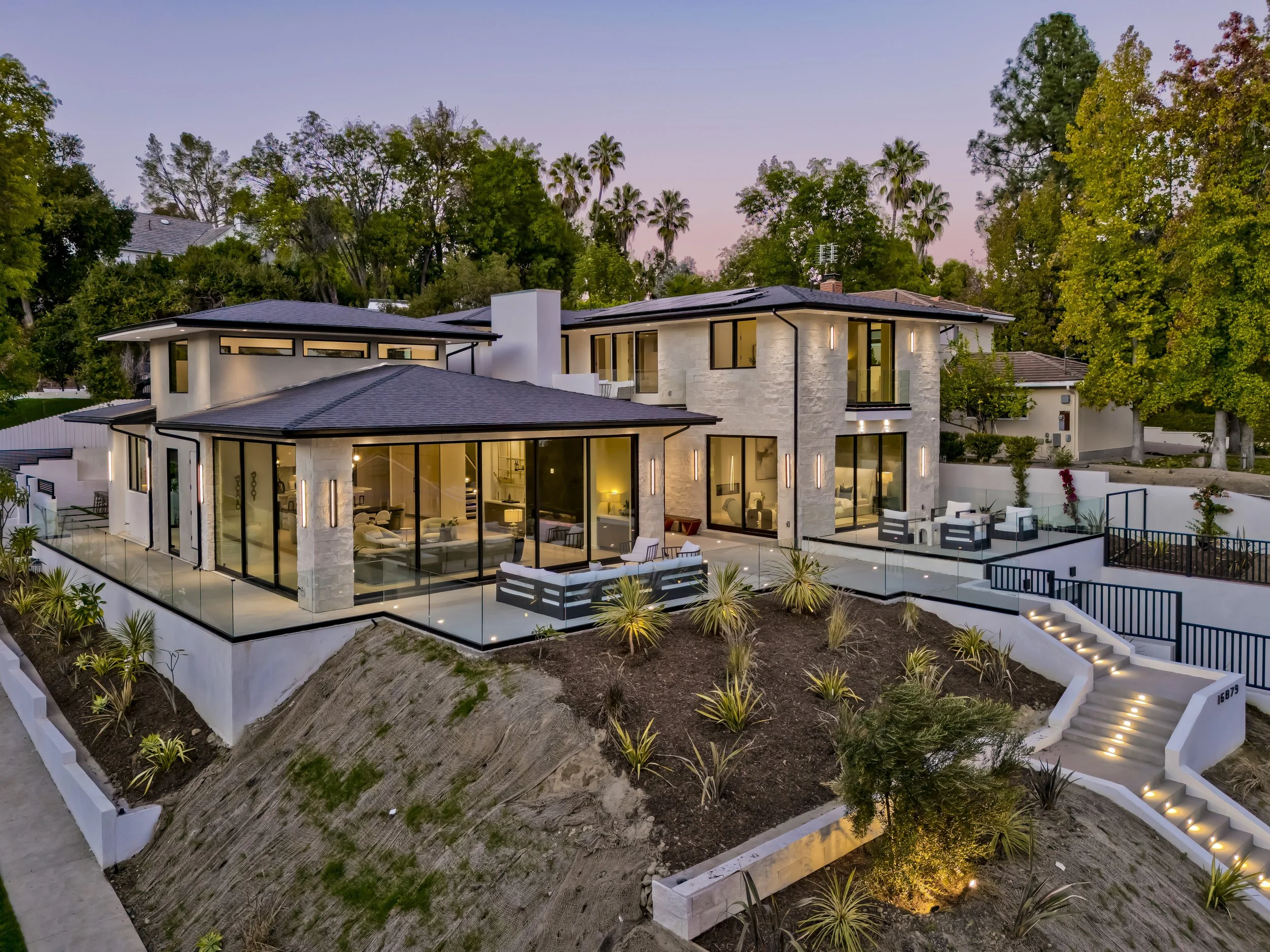 Modern custom home in Encino at dusk with stone facade, glass deck railings, and hillside landscaping
