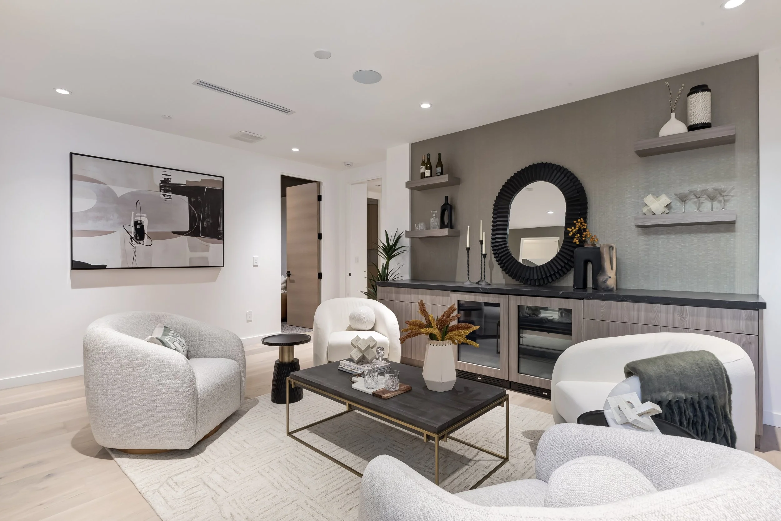 Modern living room with white and light gray furniture, a black-framed abstract art piece on the wall, wooden shelving with bottles and decorative items, a large round mirror with a black frame, and various vases and candles on a gray sideboard.