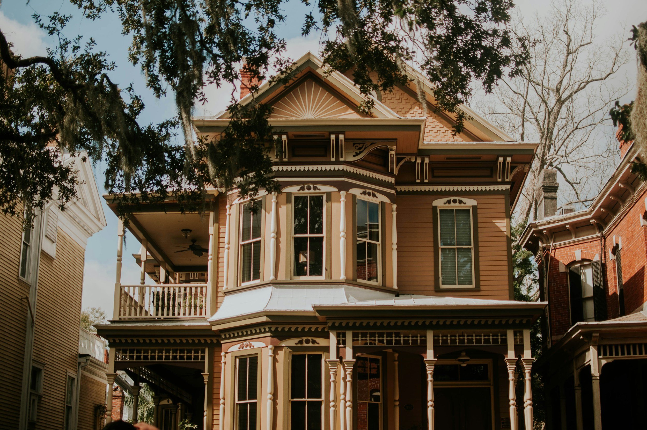 A Victorian-style house with ornate detailing, yellow and brown exterior, multiple windows, and a porch, seen through surrounding trees.