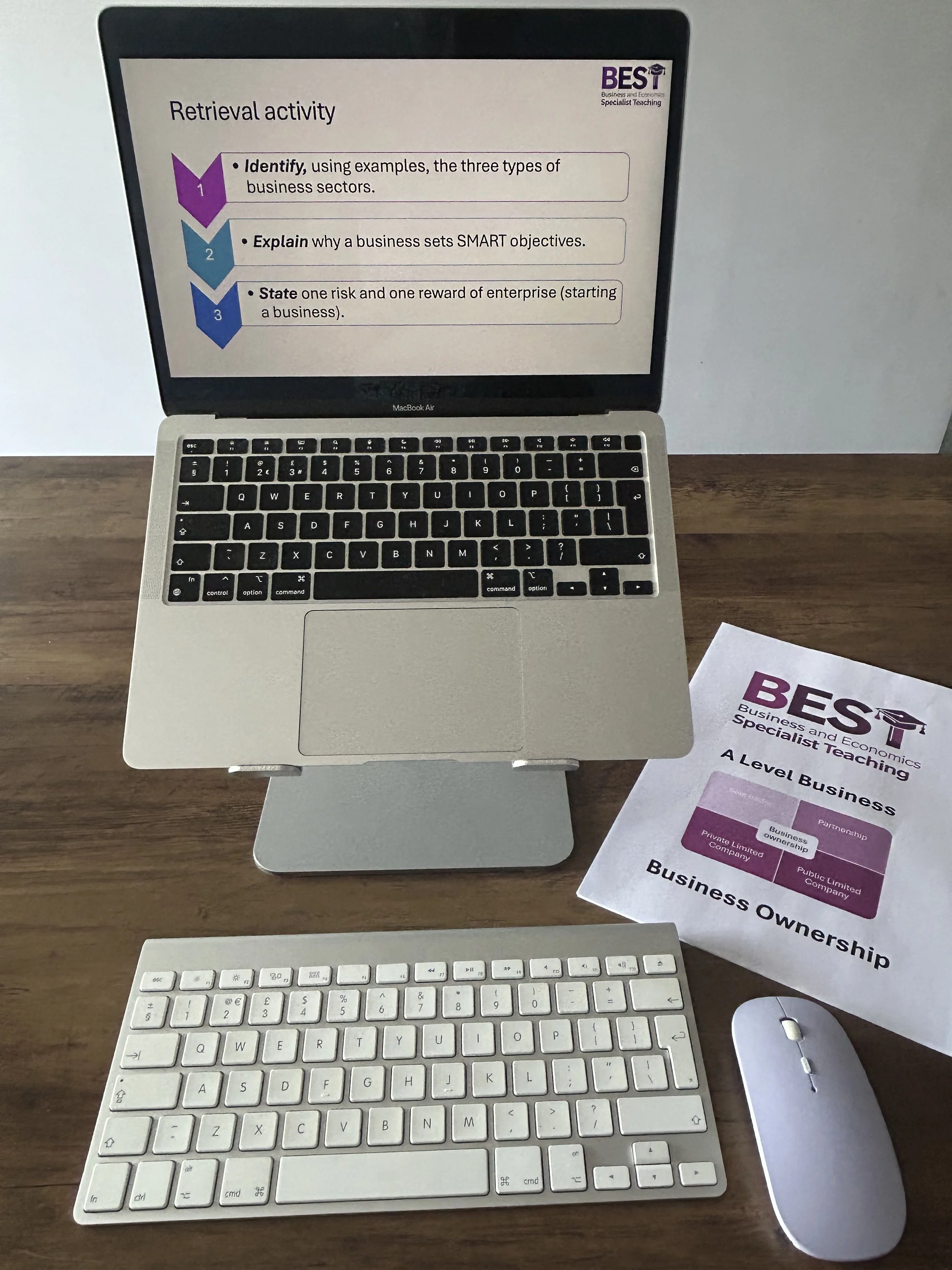 A MacBook Air laptop displaying a presentation slide about business retrieval activities, with a white wireless keyboard, a white wireless mouse, and a printed handout titled 'Business Ownership' on a wooden desk.