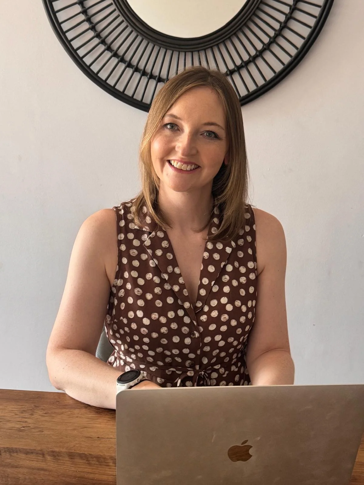 A woman with shoulder-length light brown hair, smiling, sitting at a wooden table with a silver MacBook in front of her, wearing a brown sleeveless dress with white polka dots and a smartwatch on her left wrist, behind her a round black decorative mirror on a white wall.