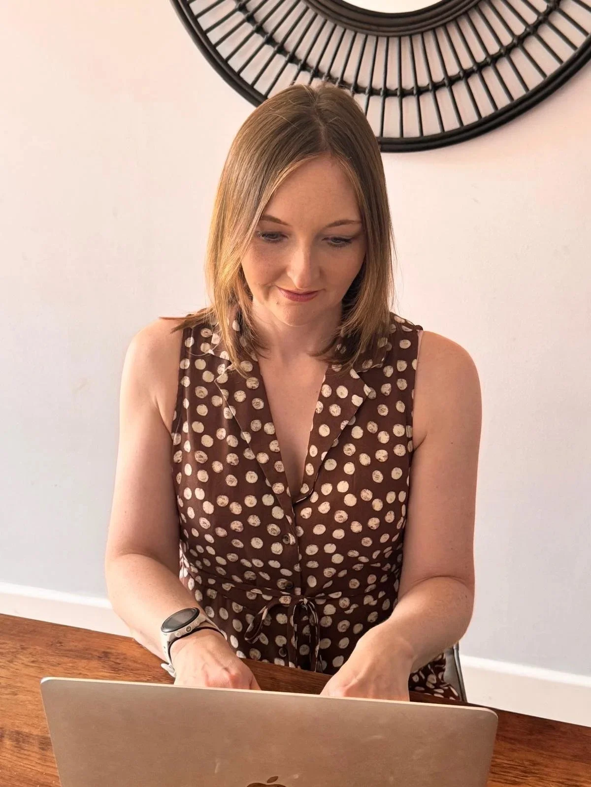 A woman with shoulder-length light brown hair wearing a brown sleeveless dress with white polka dots, sitting at a wooden table with a laptop in front of her, looking down at the laptop. There is a decorative round black wall piece behind her.