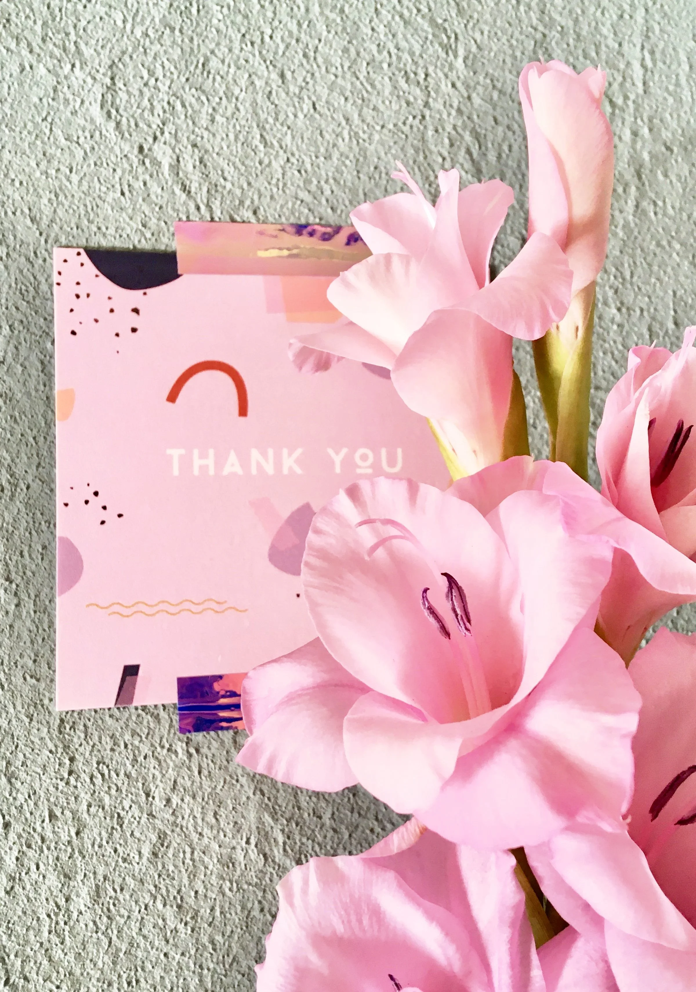 Eine rosa Dankeskarte mit Regenbögen und Muster neben pinken Blumen auf einer grün-grauen Oberfläche als Hinweis auf das nebenstehende Kontaktformular