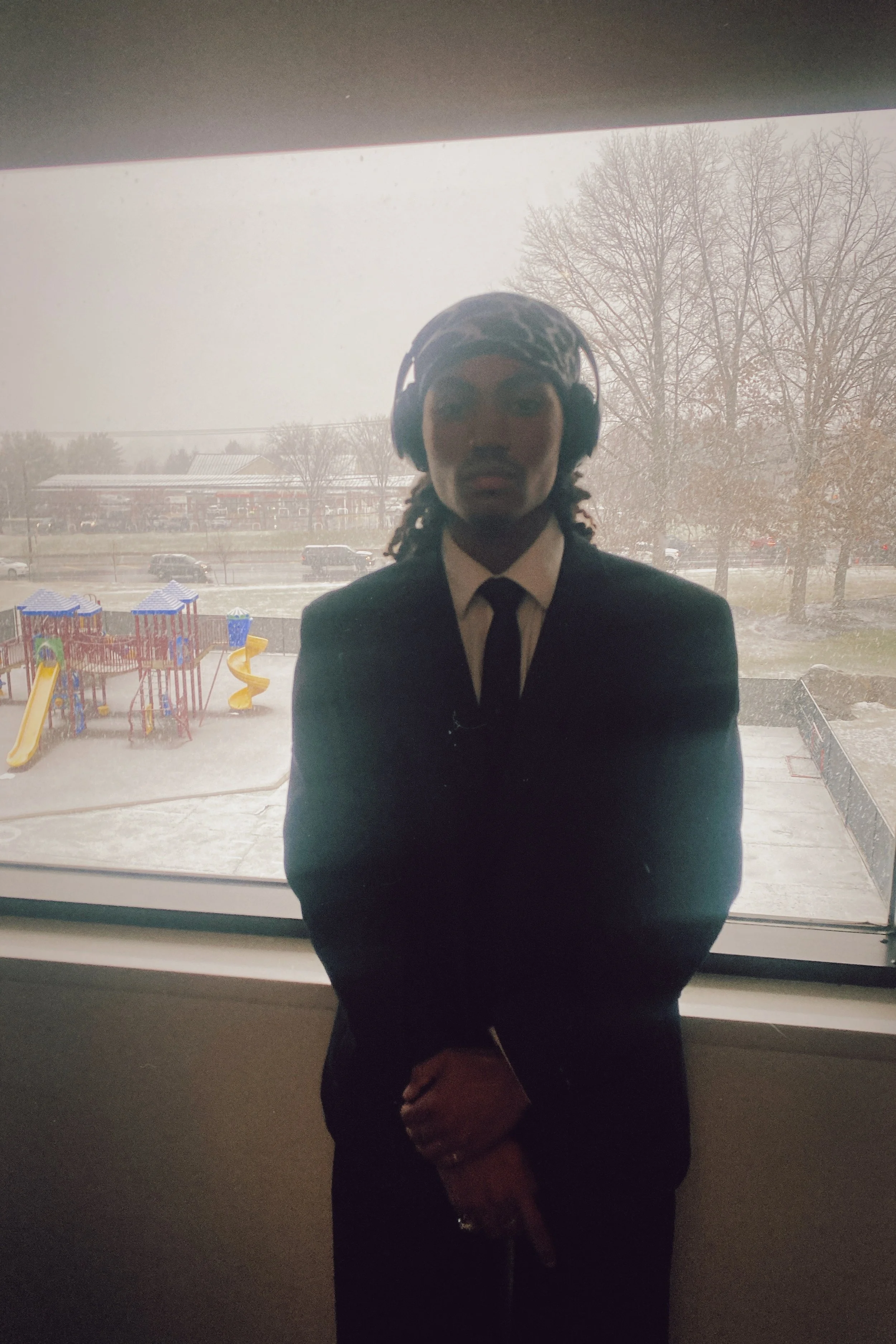 Person standing in front of a large window with a snow-covered playground and trees outside, wearing a dark suit and tie, with headphones on, and holding a bag.