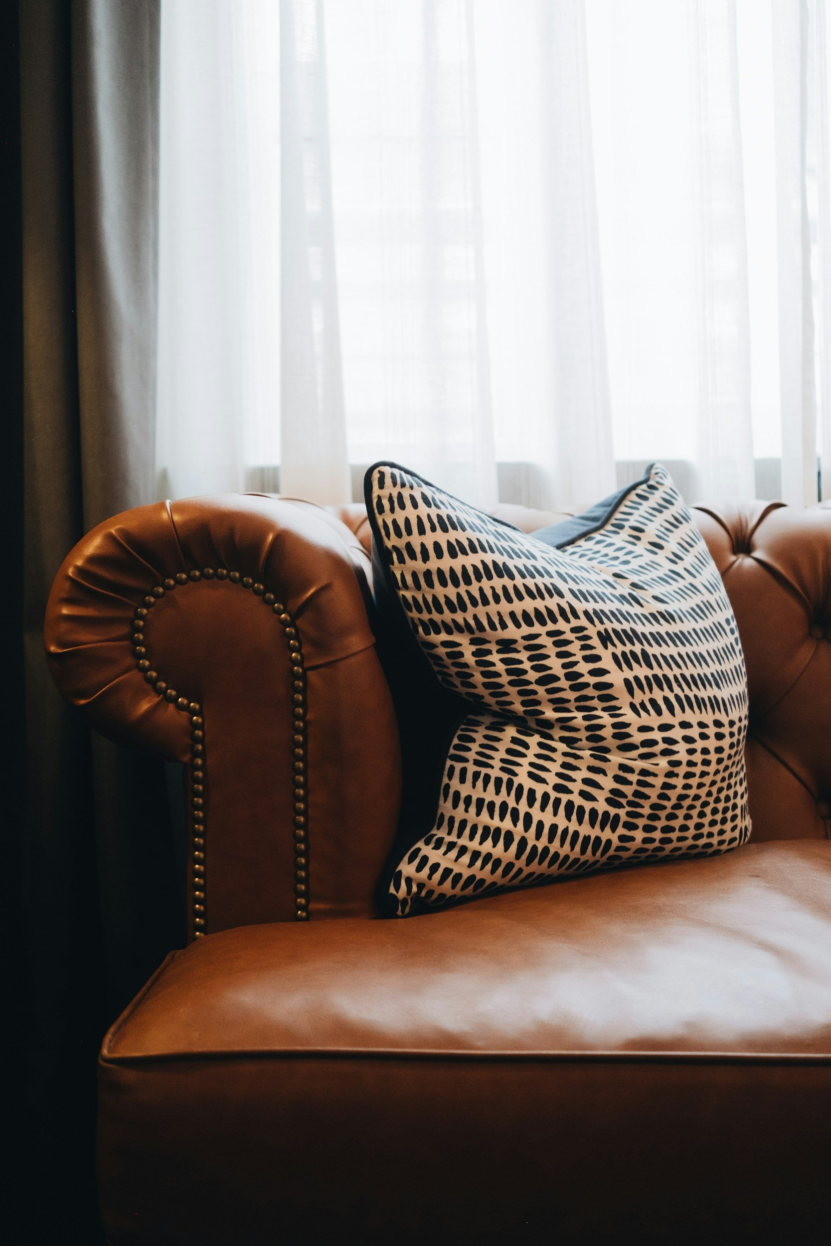Close-up of a brown leather tufted sofa with brass nailhead trim, decorated with a black and white patterned throw pillow, against a window with sheer white curtains.