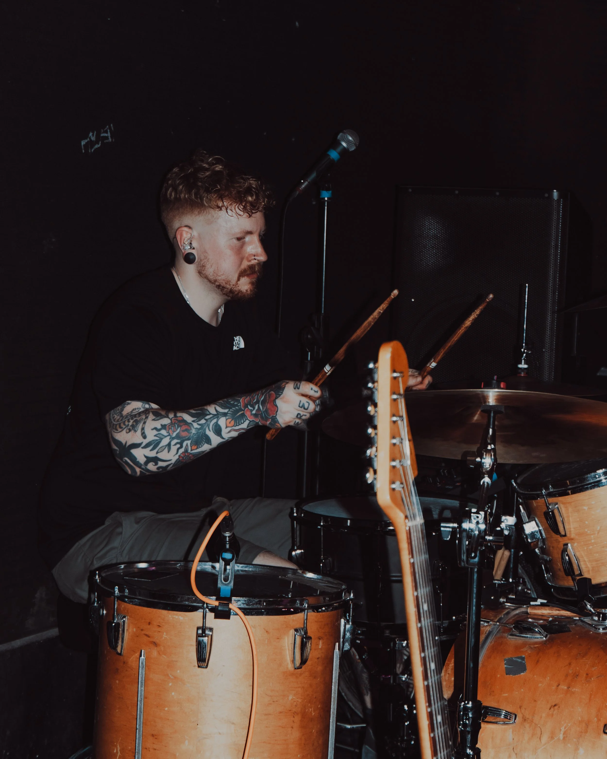 A white man with ginger hair and ginger facial hair plays drums at a live gig
