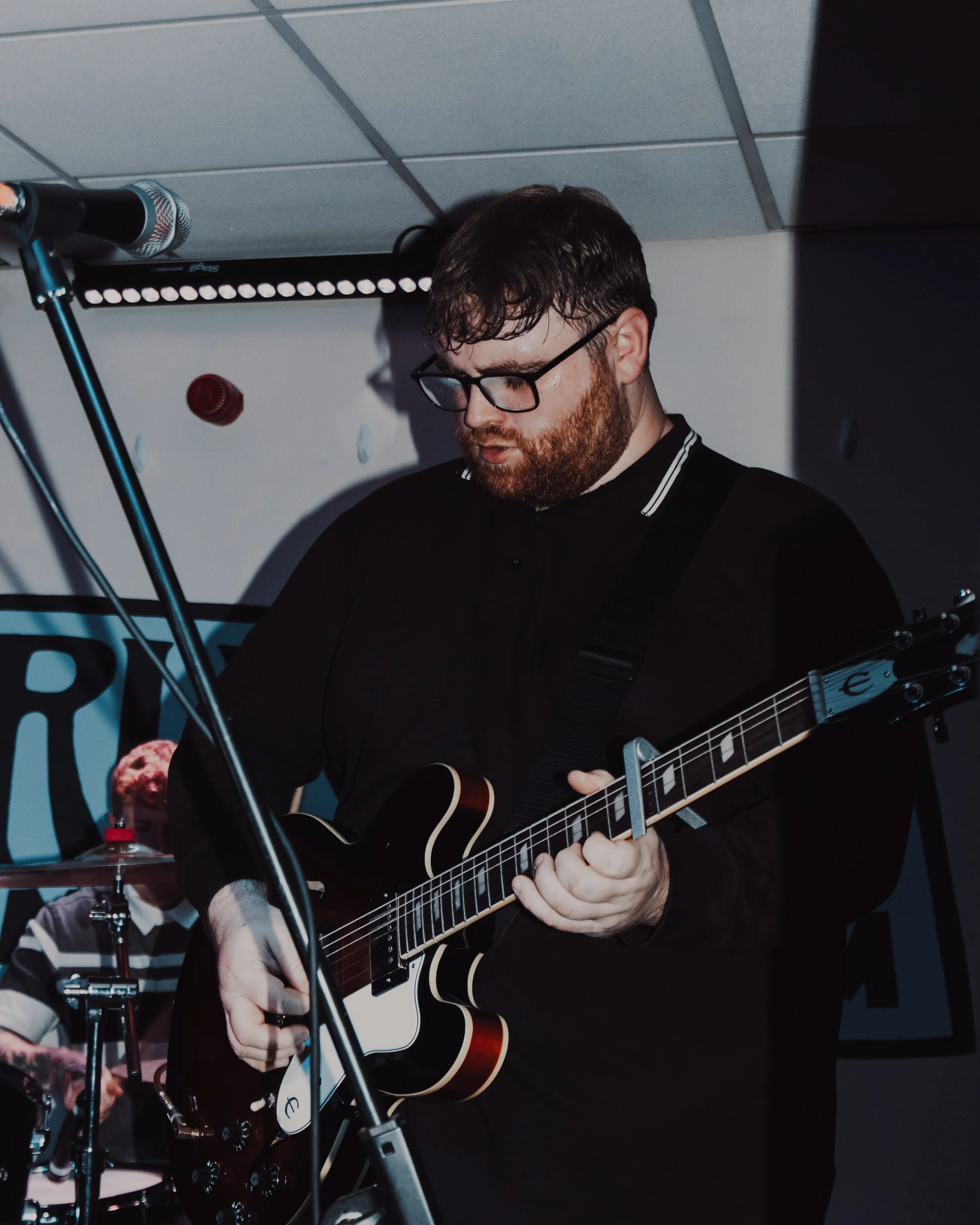 A tall, stocky white man with brown hair and a brown beard plays electric guitar at a live gig