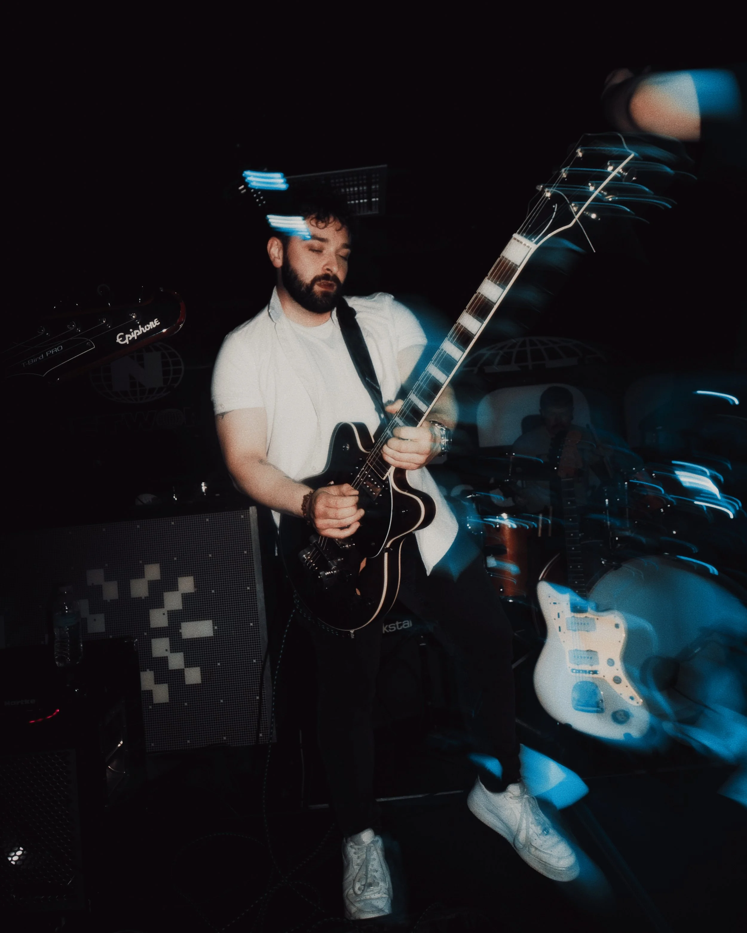 A short white male with black hair and a black beard plays lead electric guitar at a gig