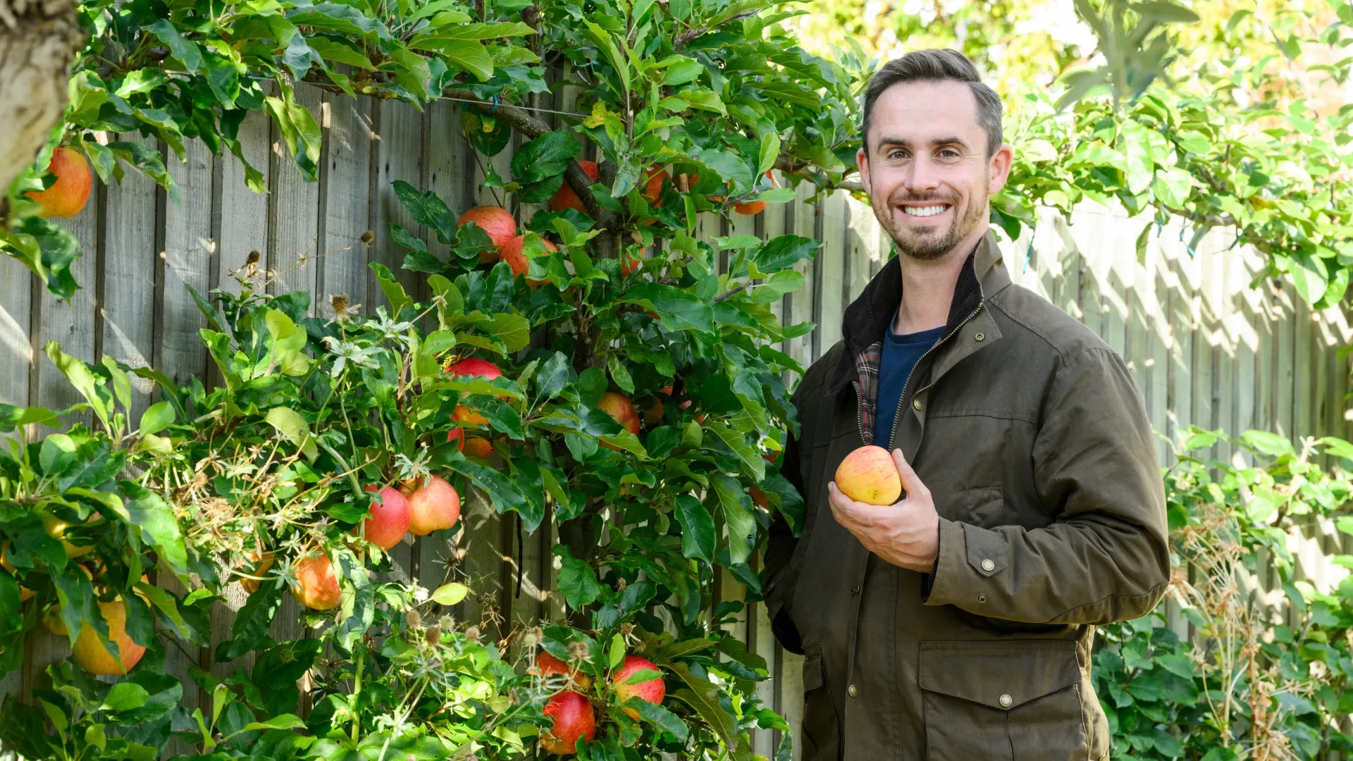 Michael Griffiths in a brown jacket holding an apple standing in a garden next to an apple tree with red apples, in a backyard with a wooden fence and green foliage.
