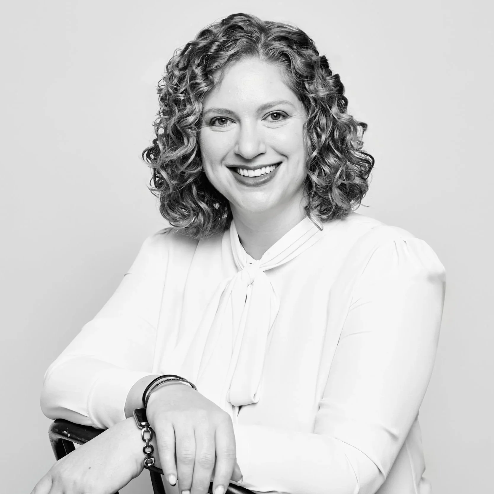 Black and white portrait of a woman with curly hair smiling, wearing a light-colored blouse and bracelets, posing with her arms crossed.