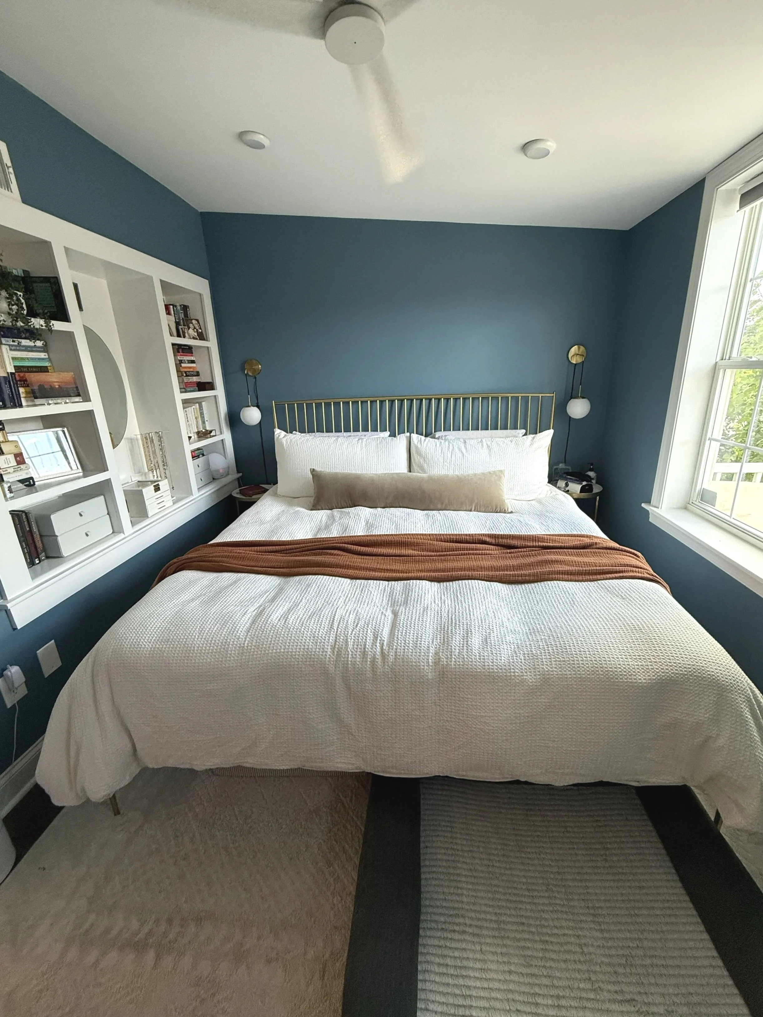 Bedroom with a bed, white bedding, gold headboard, blue walls, large window, bookshelves, wall-mounted lamps, and a colorful area rug.