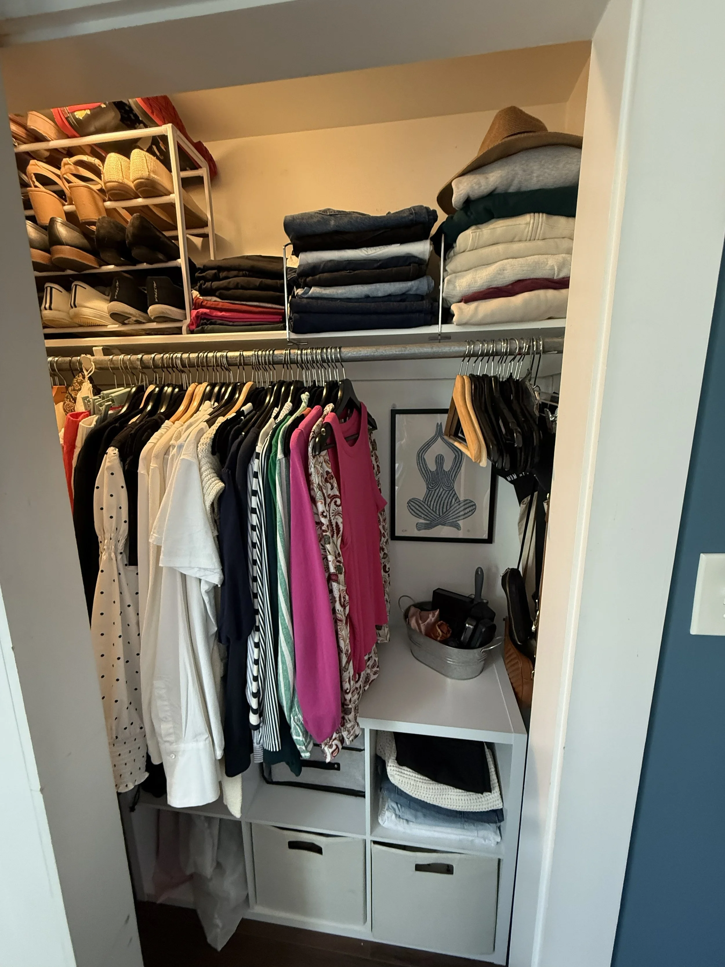 A walk-in closet with shelves and hanging clothes, including shirts and dresses, and neatly folded pants and sweaters. Shoes are stored on a shelf, and there is a framed drawing of a figure sitting cross-legged on the wall.